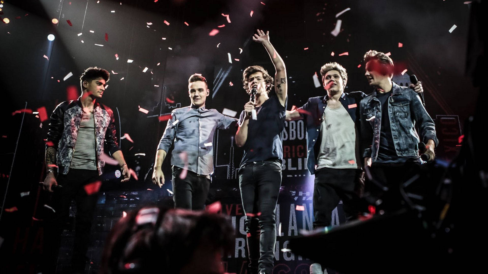 One Direction 舞台熱唱 壁紙 — 五名男性樂隊成員在黑暗的舞台上，在聚光燈下表演，紅色紙屑在他們周圍飄落。