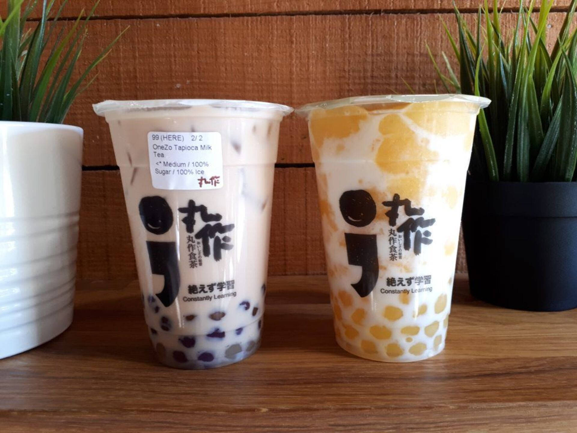 OneZo 珍珠奶茶 壁紙 — 兩杯珍珠奶茶，含有木薯珍珠和芒果珍珠，放在一張木桌上，兩旁有盆栽植物。