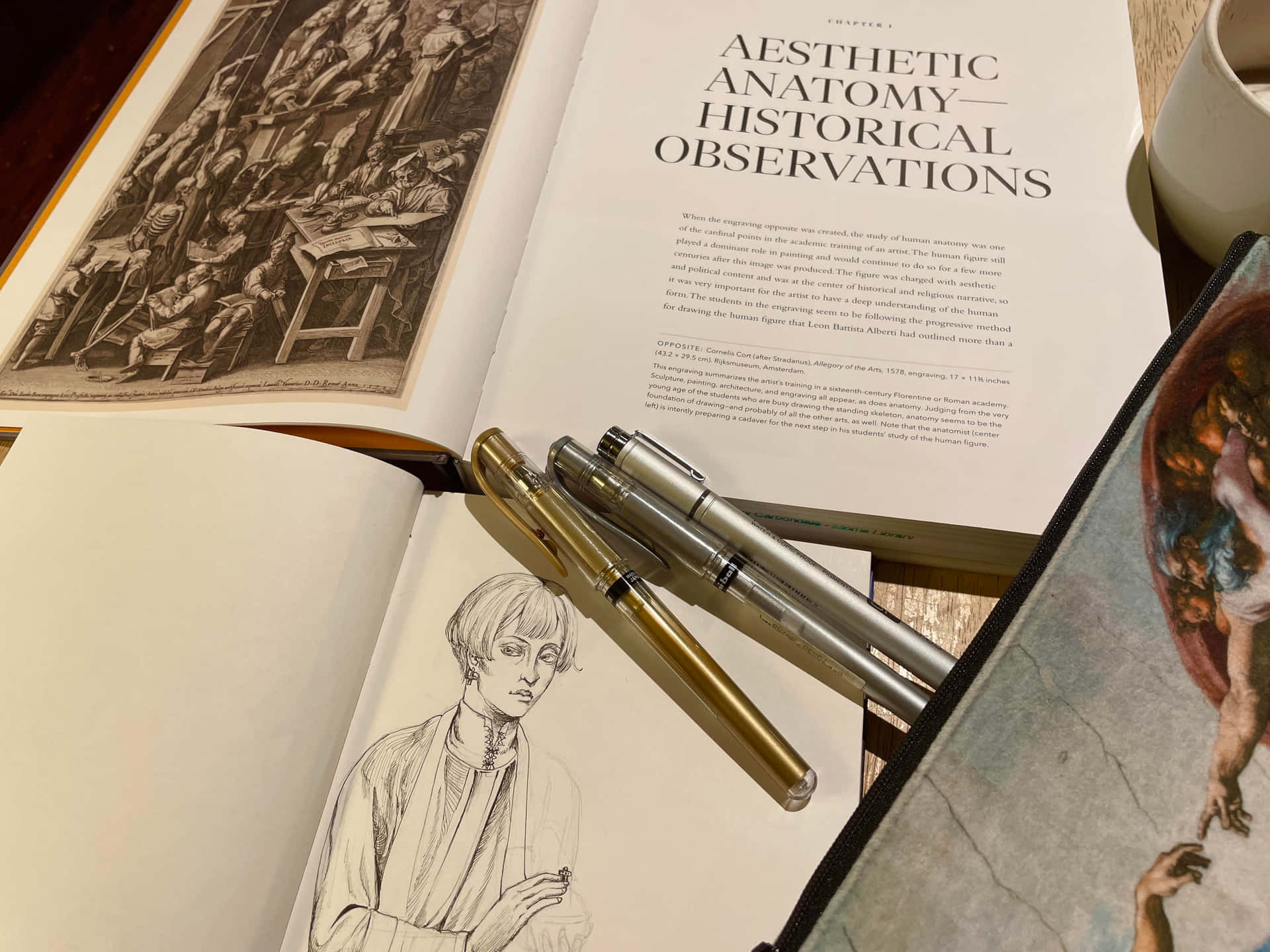  壁紙 — 一本打開的藝術書，內有歷史插圖和一幅女性素描，旁邊有兩支筆放在木桌上。