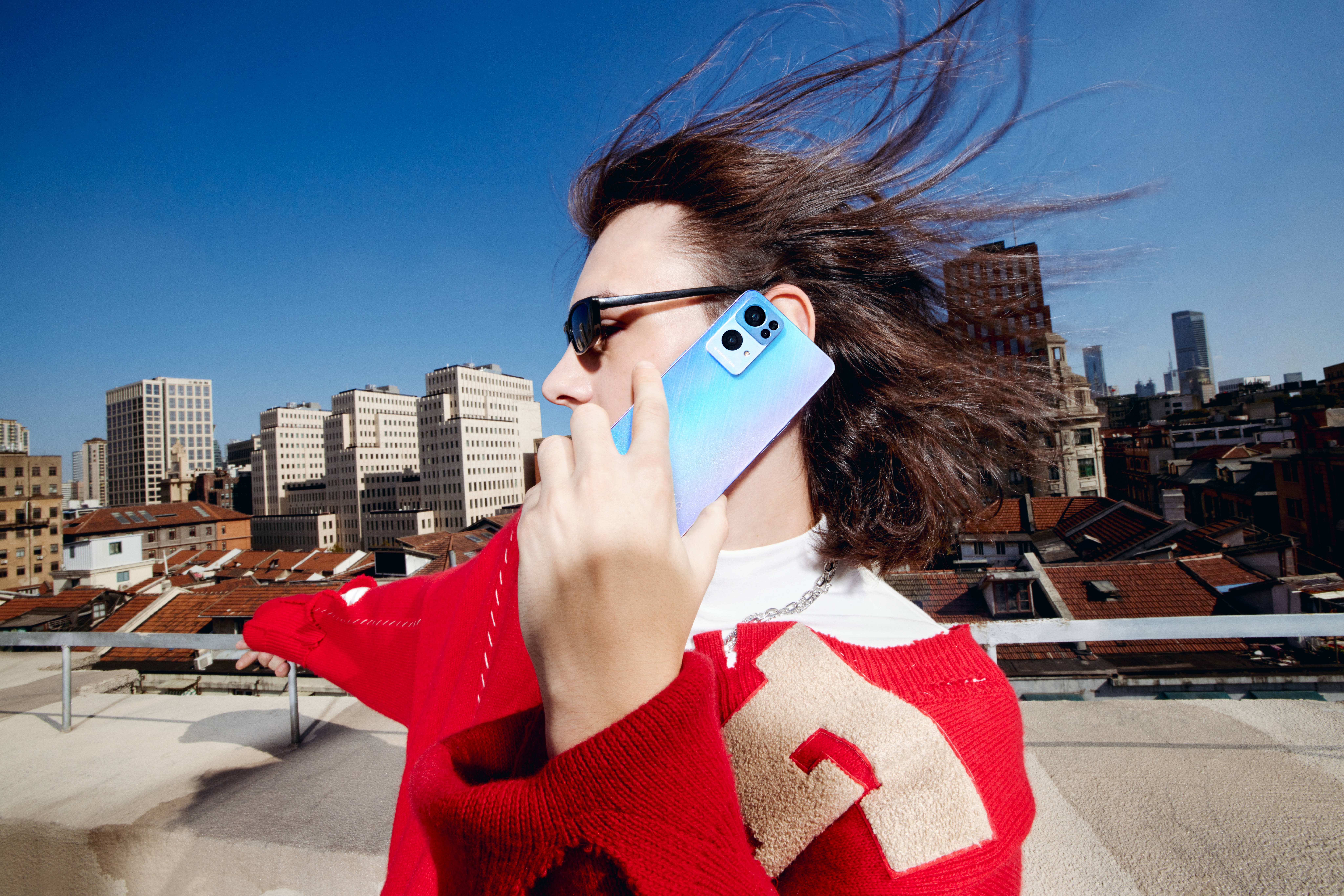 通話中的Oppo Reno7 壁紙 — 一位身穿紅色毛衣的女性在都市屋頂上使用藍色電話通話，風吹動她的頭髮，背景是清澈的藍天。