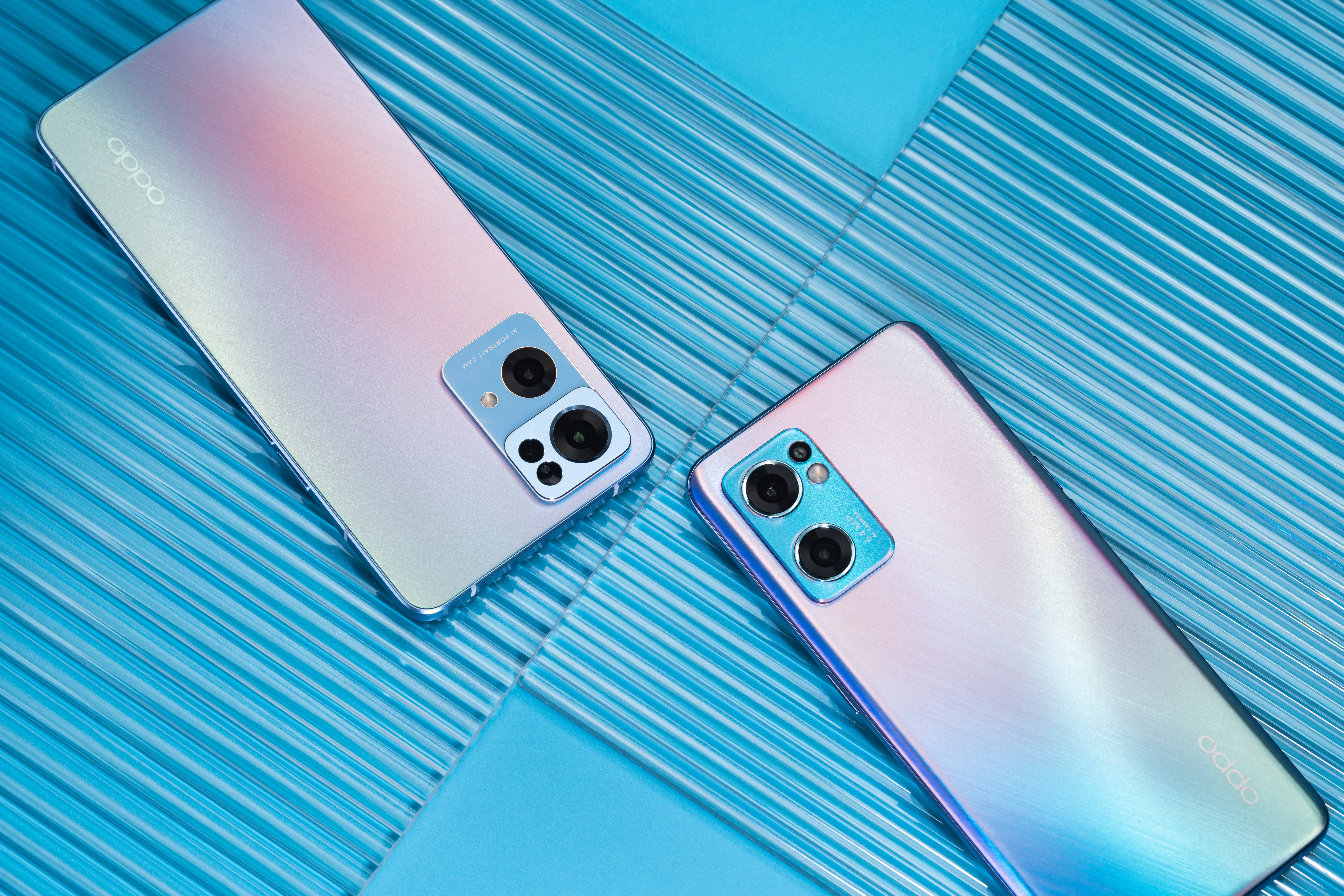 Oppo Reno7 系列 壁紙 — 兩部帶有彩虹色漸變背面的OPPO智能手機對角放置在帶紋理的藍色表面上。