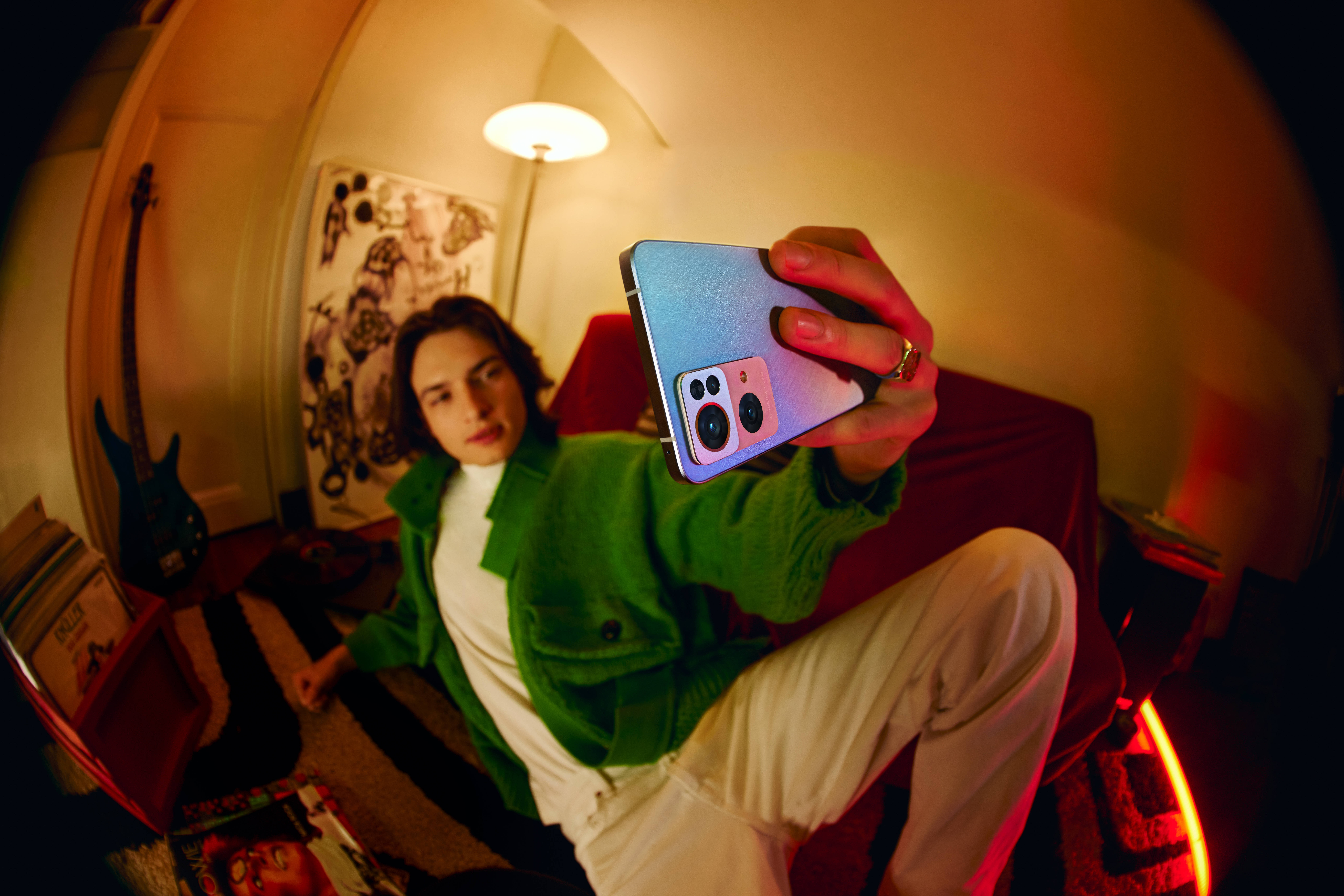 Oppo Reno7自拍 壁紙 — 一個穿著綠色外套的年輕人在一個溫暖光線、雜亂的房間裡用彩色手機拍攝魚眼自拍。