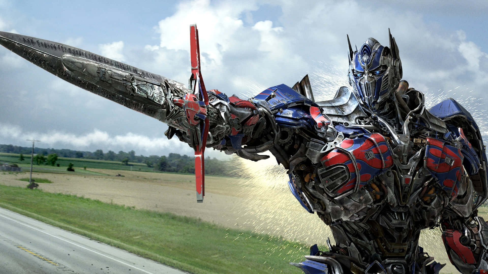  壁紙 — Optimus Prime 以機械人形態，手持帶有紅色點綴的巨大劍，在鄉村道路上強勢前行。