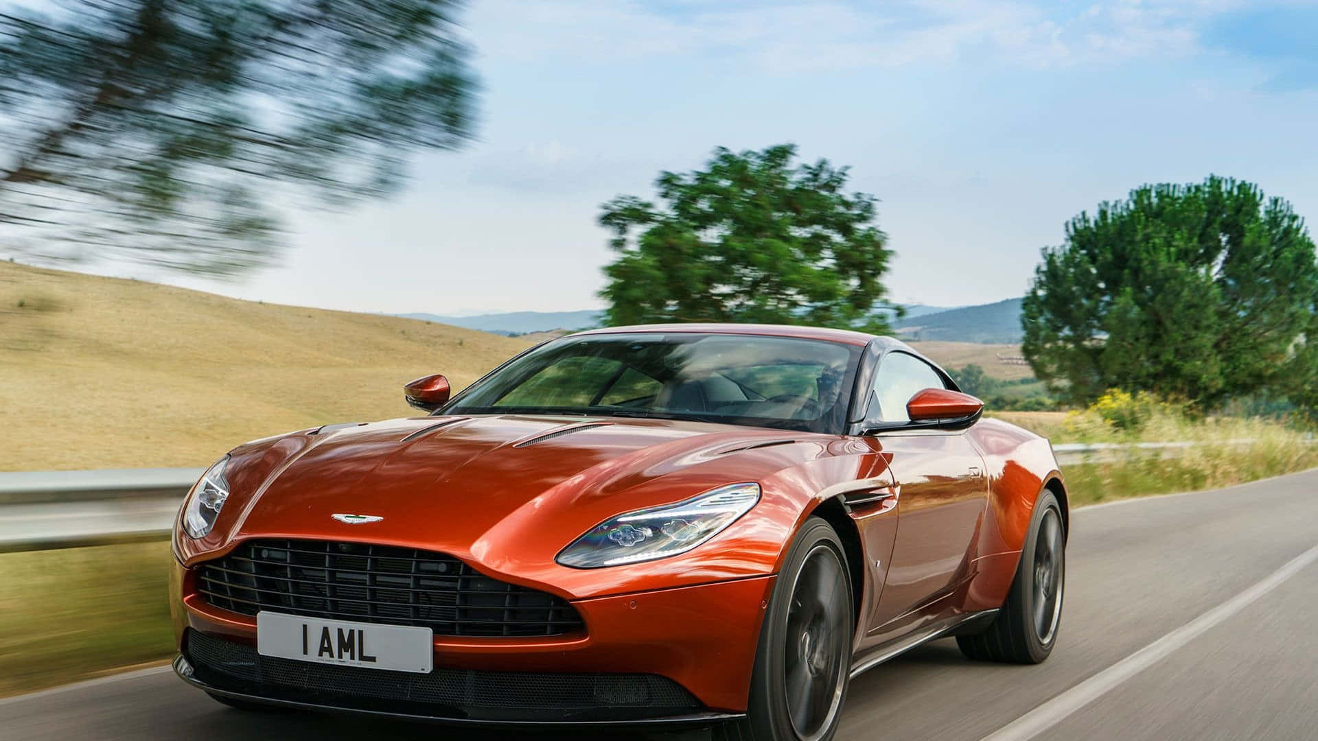 Aston Martin Db11 桌布 壁紙 — 一輛流線型橙色 Aston Martin 在蜿蜒的鄉村道路上飛馳，背景是模糊的綠樹和金色山丘，上方是明亮的藍天。