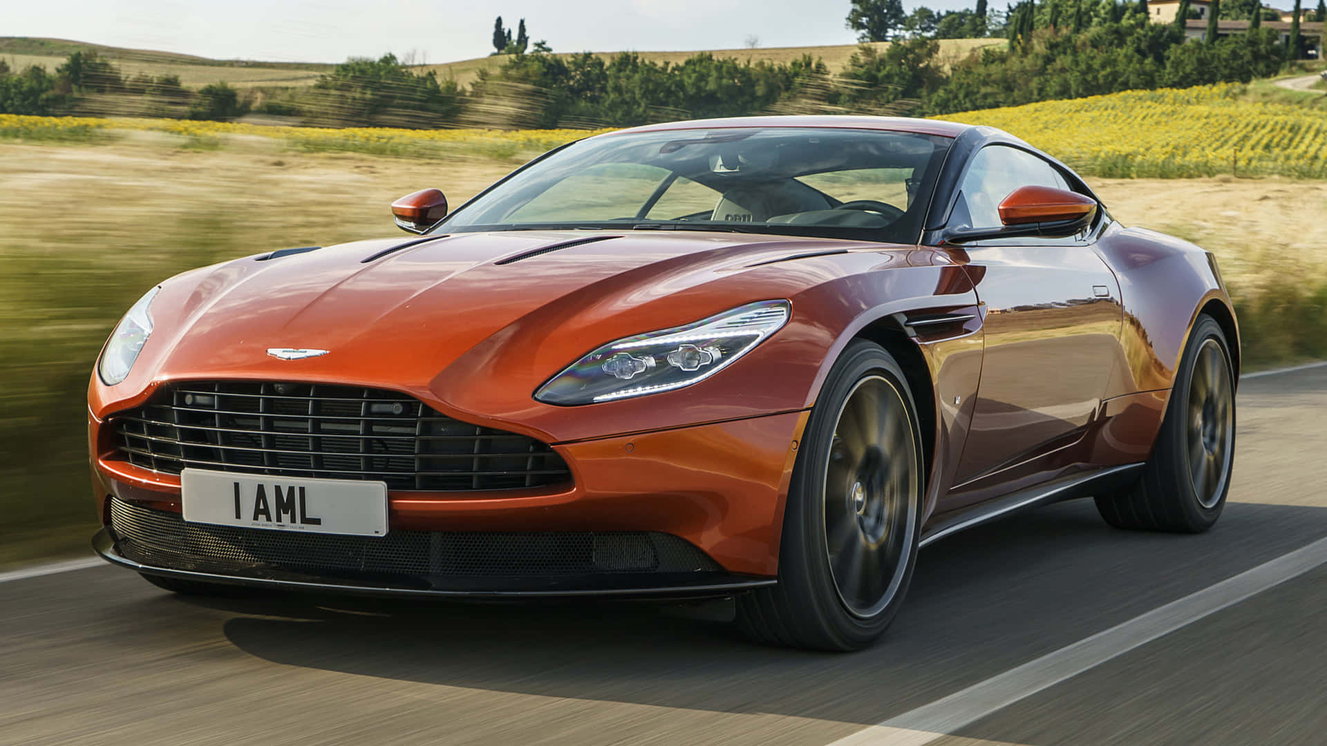 Aston Martin Db11 桌布 壁紙 — 一輛流線型的橙色Aston Martin跑車在陽光明媚的鄉村道路上疾馳，背景是模糊的金色田野和樹木。