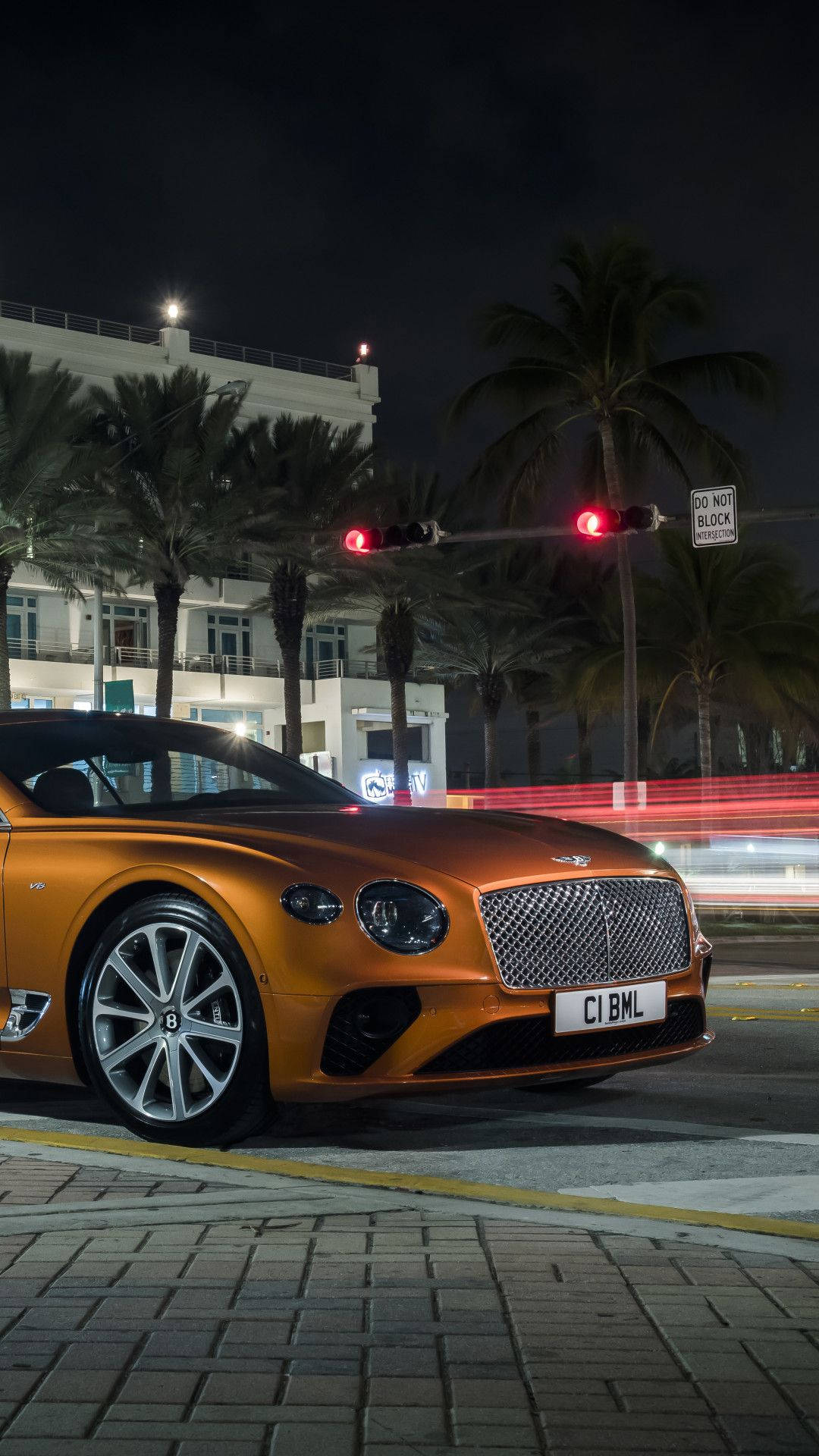 橙色 Bentley 豪華車夜間停泊在城市的街道上，有棕櫚樹和發光的交通燈。