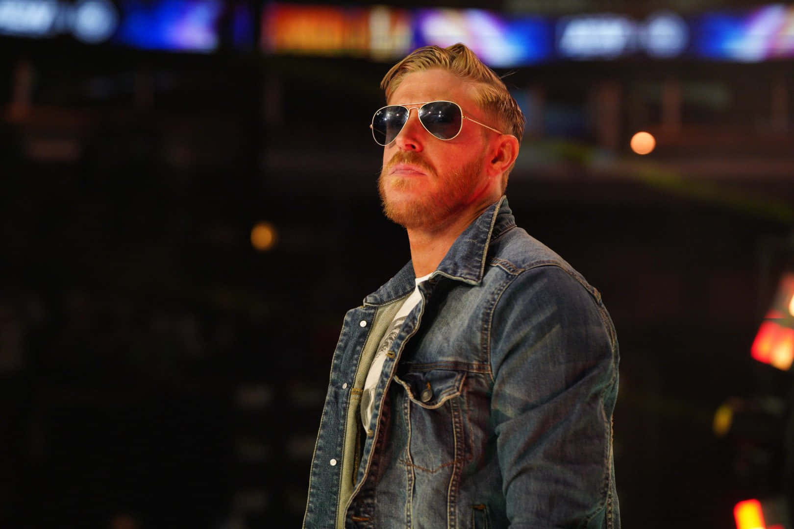 Orange Cassidy - AEW 摔角界的慵懶強者 壁紙 — 一位留鬍鬚的男子戴著飛行員太陽眼鏡，自信地站在燈光昏暗的競技場中，身後有色彩繽紛的模糊燈光。
