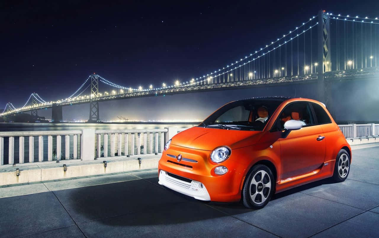 Fiat 500l 桌布 壁紙 — 一輛鮮橙色的Fiat 500在夜間停泊在發光的吊橋旁邊，橋下是水面。