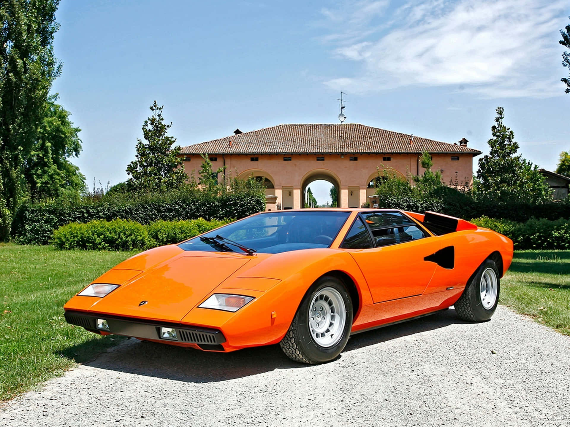 Lamborghini Countach 桌布 壁紙 — 一輛鮮橙色的林寶堅尼Countach停泊在碎石車道上，前方是一座帶拱形入口和茂密綠植的灰泥別墅，背景是明亮的藍天。