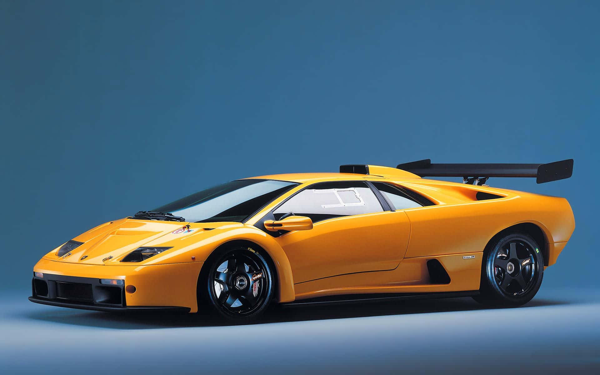 Lamborghini Diablo 桌布 壁紙 — 一輛充滿活力的橙色林寶堅尼 Diablo SV 跑車，帶有黑色飾邊和大型後尾翼，在純藍色工作室背景前展示。