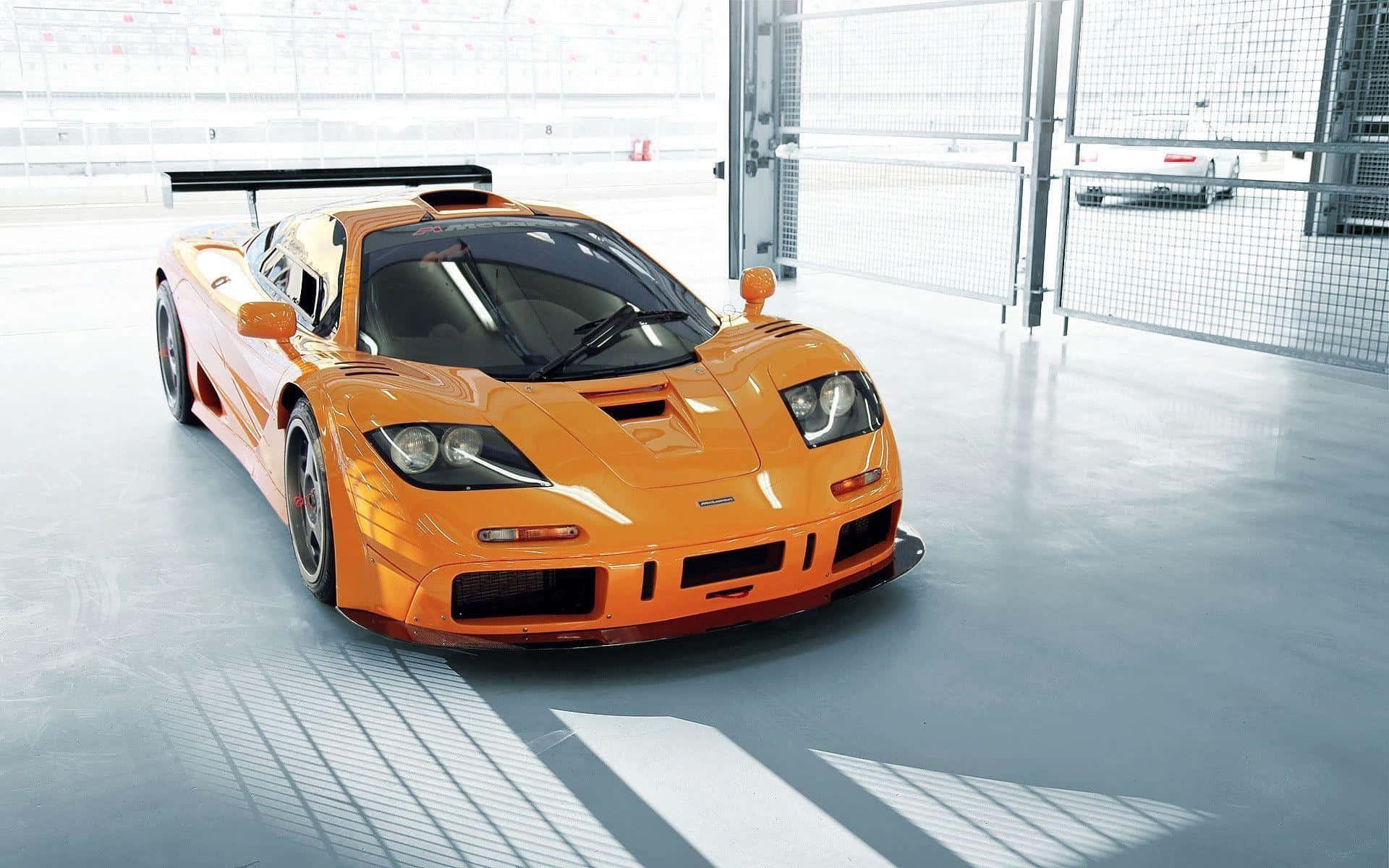 Mclaren F1 桌布 壁紙 — 一輛鮮豔的橙色McLaren F1超級跑車在明亮燈光下，於簡約時尚的車庫中閃耀，地板上有網格狀陰影。