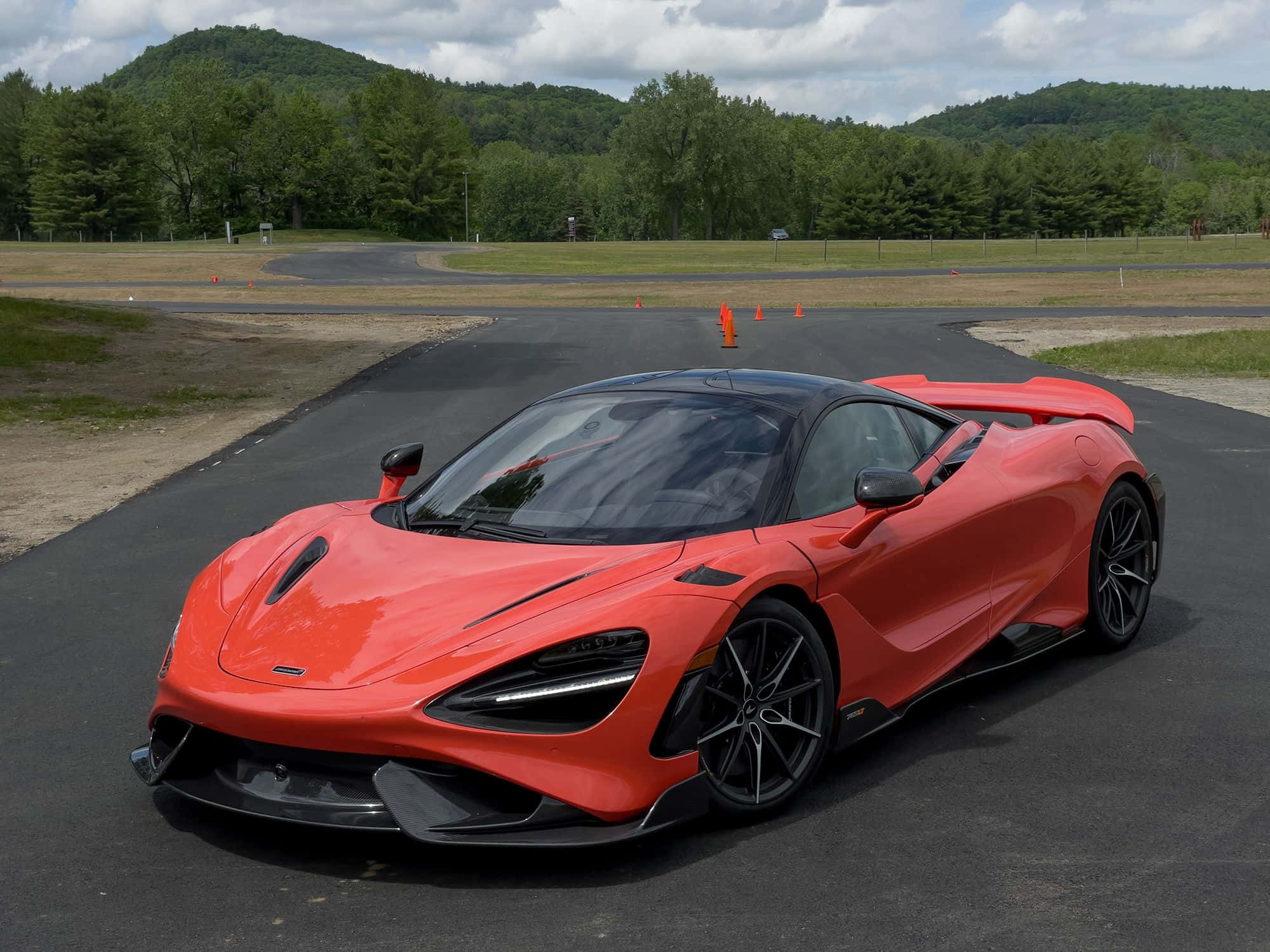 Mclaren 765lt 桌布 壁紙 — 一輛充滿活力的橙色McLaren跑車在蜿蜒穿過森林山坡的賽道上閃閃發光，天空多雲。