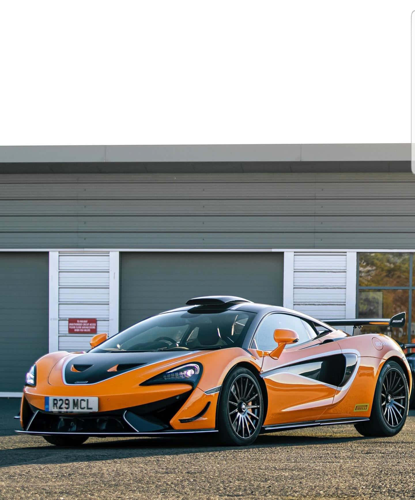 Mclaren 620r 桌布 壁紙 — 一輛充滿活力的橙色McLaren跑車，帶有黑色飾邊，停在現代工業建築前的碎石地上。