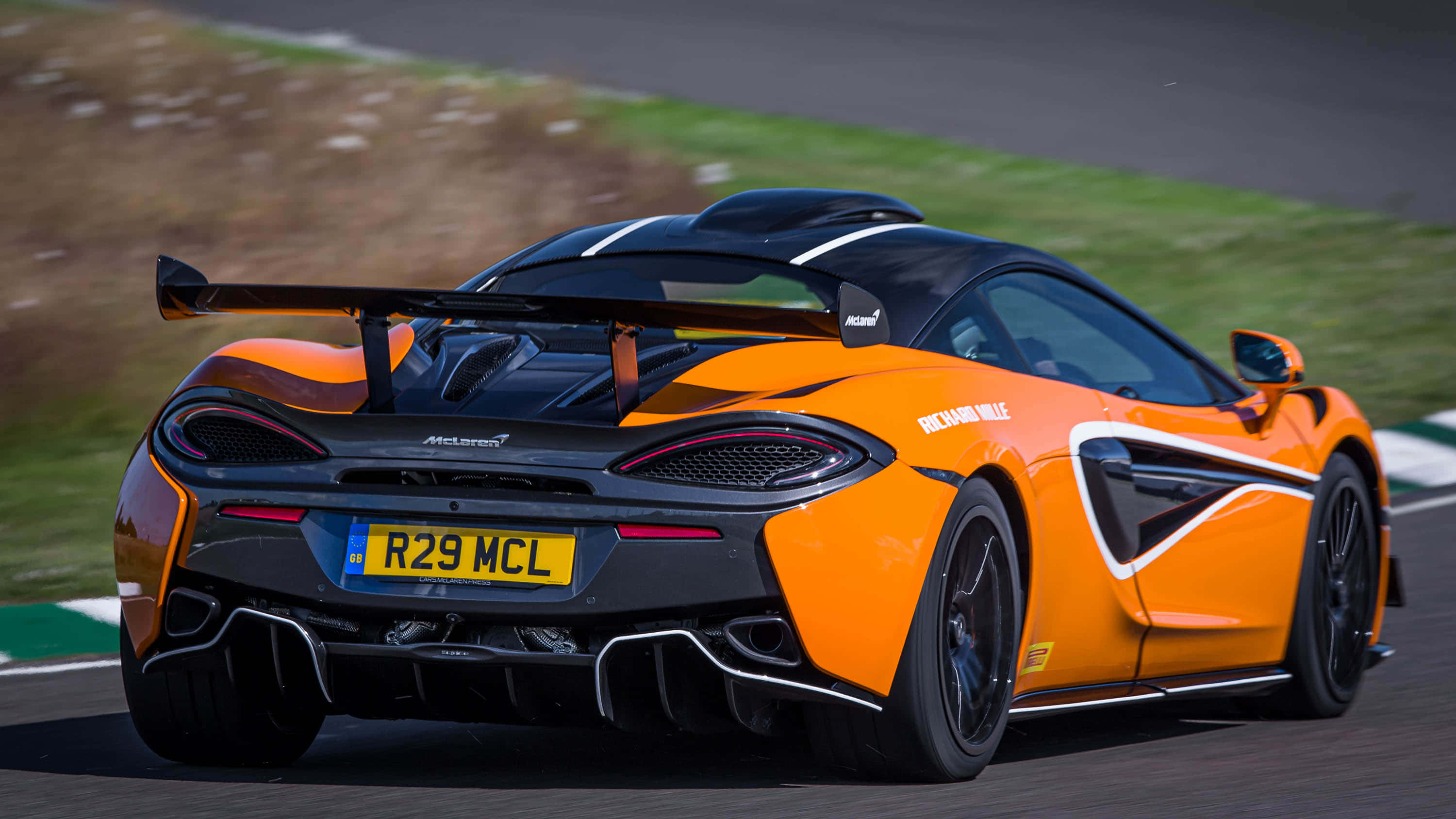 Mclaren 620r 桌布 壁紙 — 一輛充滿活力的橙色McLaren跑車帶有黑色點綴，在賽道上疾馳，背景是模糊的綠色草地和瀝青。