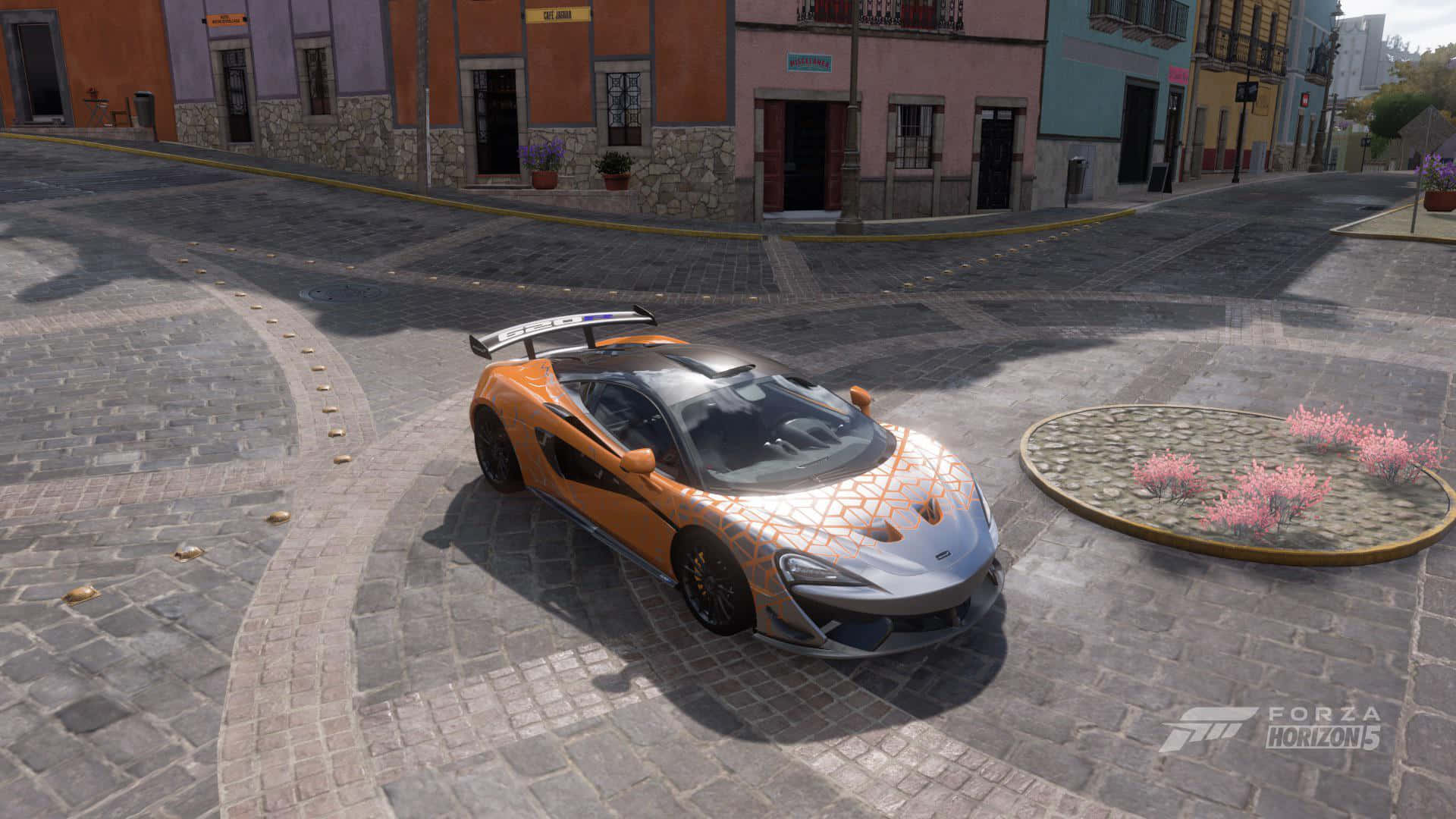 Mclaren 620r 桌布 壁紙 — 一輛充滿活力的橙色和銀色麥拿侖跑車停泊在鵝卵石廣場上，周圍是色彩繽紛的殖民時期建築。