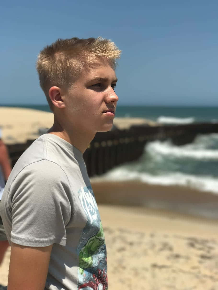 可愛的《Outer Banks》男孩 壁紙 — 一個小男孩在陽光明媚的海灘上側身站立，凝視著海浪拍打木製碼頭的海洋。