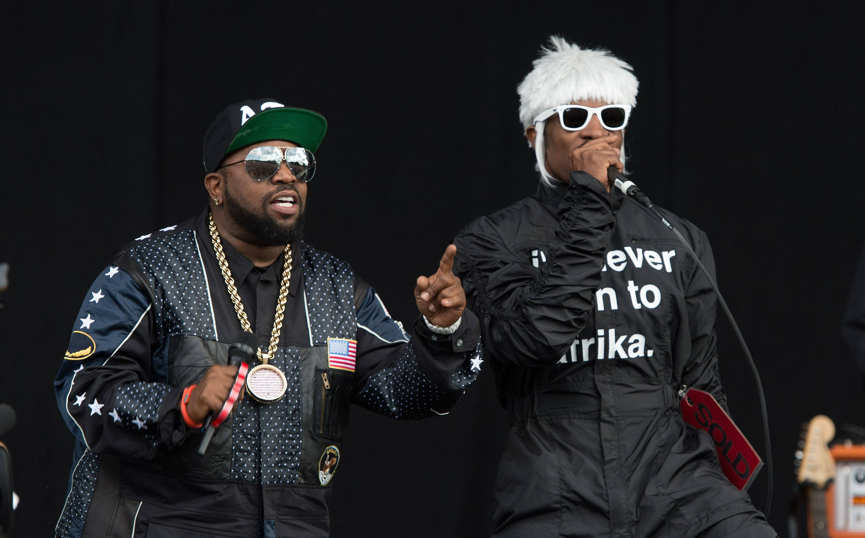 Outkast 大波伊與安德烈3000饒舌表演 壁紙 — 兩位說唱歌手在舞台上充滿活力地表演，一位身穿星星圖案外套和金色鏈條，另一位戴著白色假髮和太陽眼鏡。