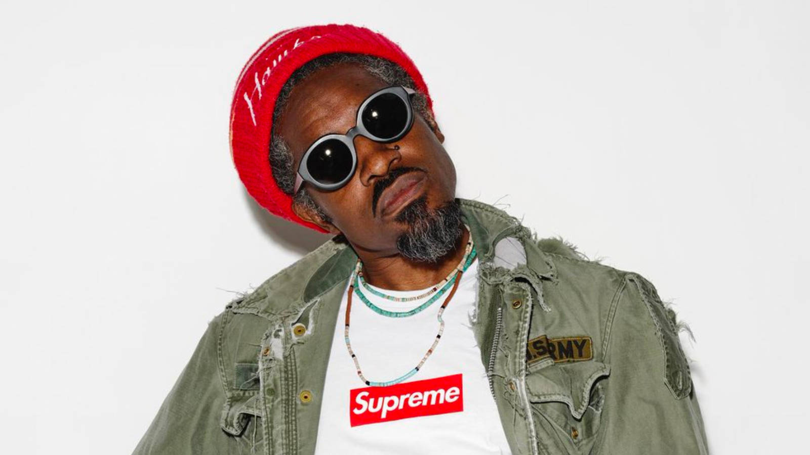男士戴紅色冷帽太陽眼鏡Supreme T恤綠色外套在白色背景前擺姿勢