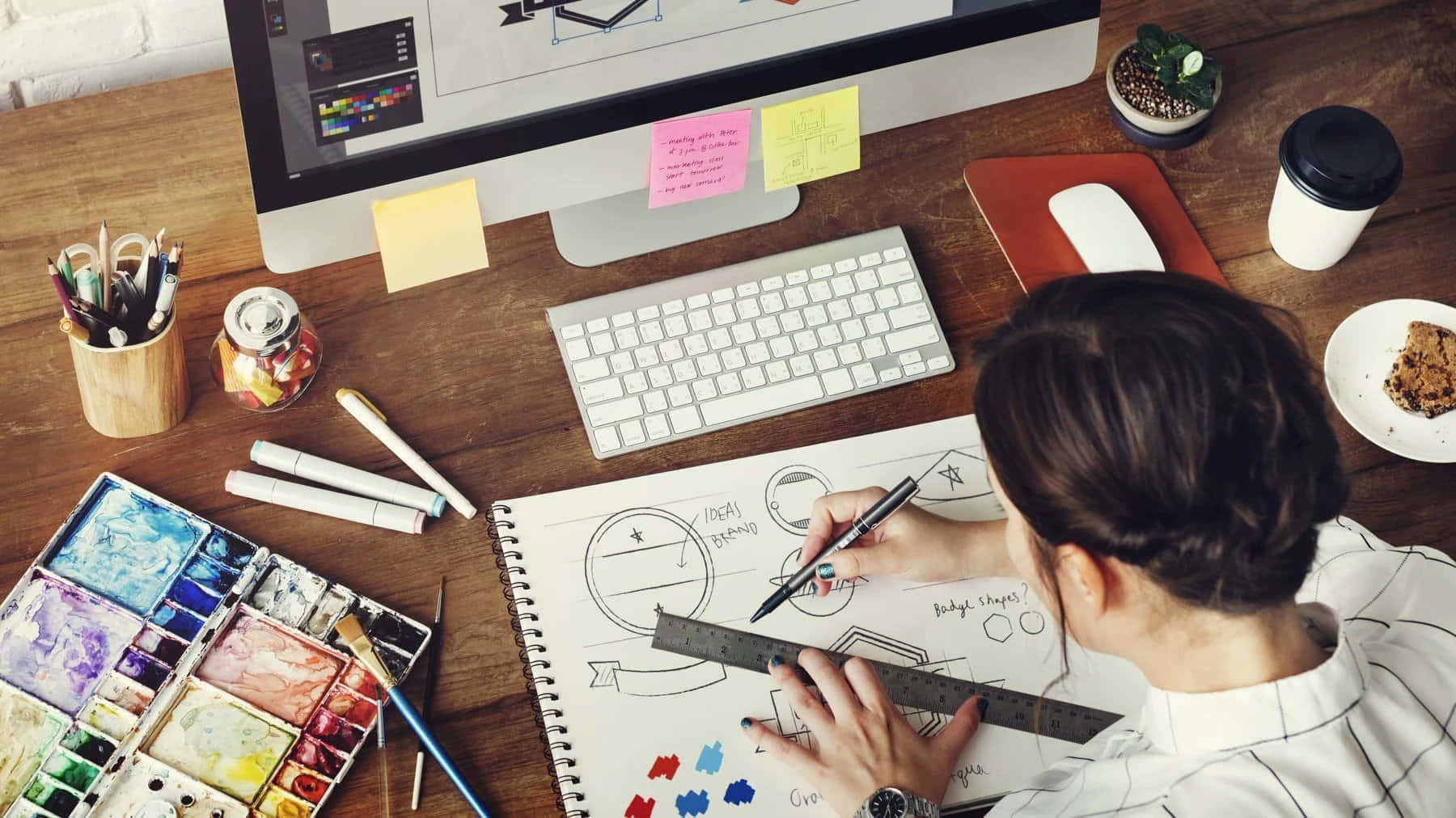 graphic design 桌布 壁紙 — Overhead view of a designer sketching with ruler and pen at a wooden desk cluttered with art supplies and a computer.