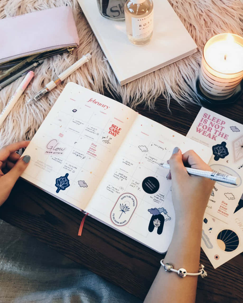  壁紙 — Overhead view of hands writing in a February bullet journal on a fluffy rug surrounded by pens stickers and a lit candle.