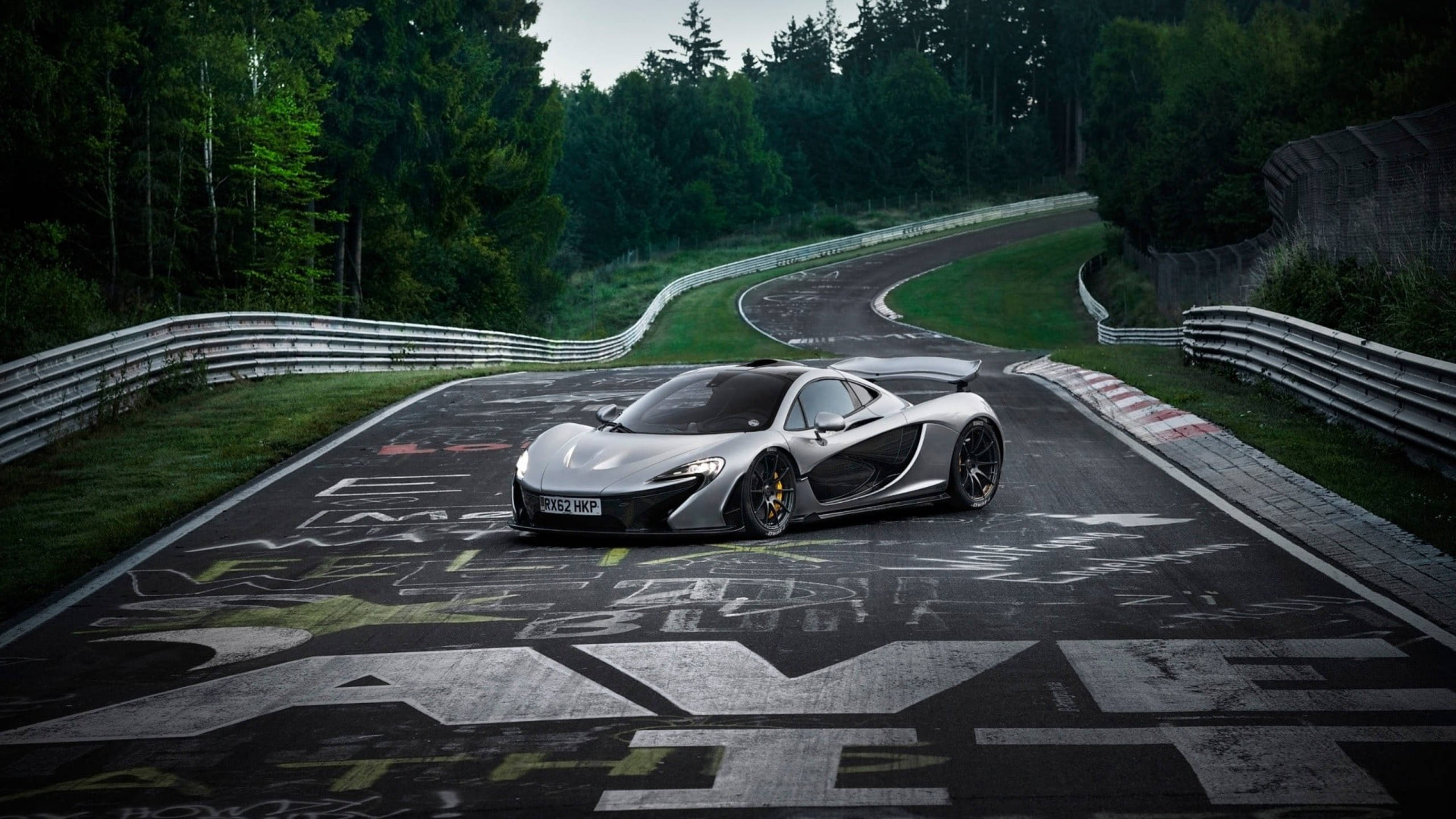 麥拉倫P1賽車 壁紙 — 一架流線型的銀色McLaren P1跑車在蜿蜒的瀝青賽道上行駛，周圍是茂密的綠色森林。