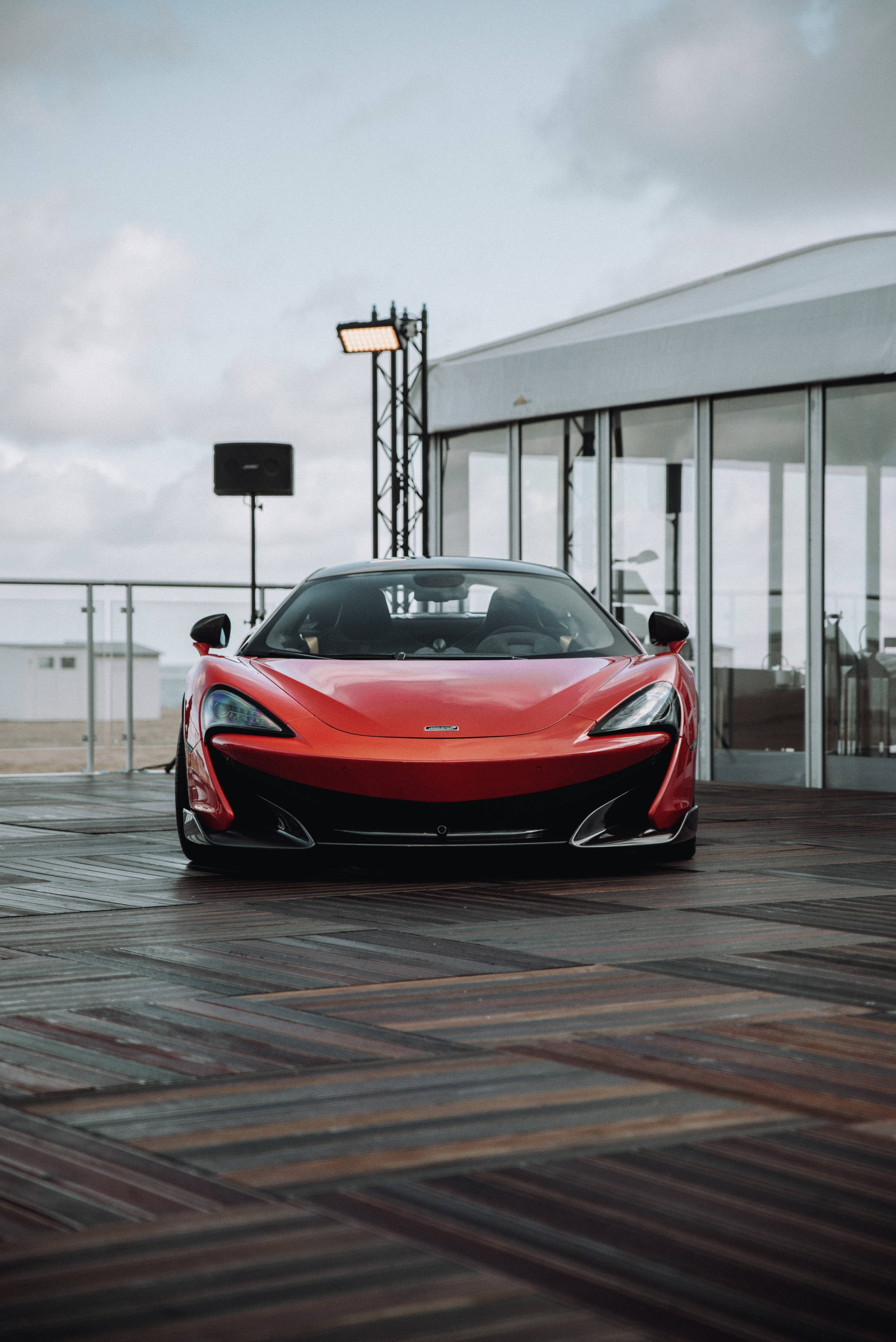 紅黑配色麥拉倫P1 壁紙 — 一輛鮮紅色的McLaren跑車停在現代玻璃建築附近的木質甲板上，天空多雲。