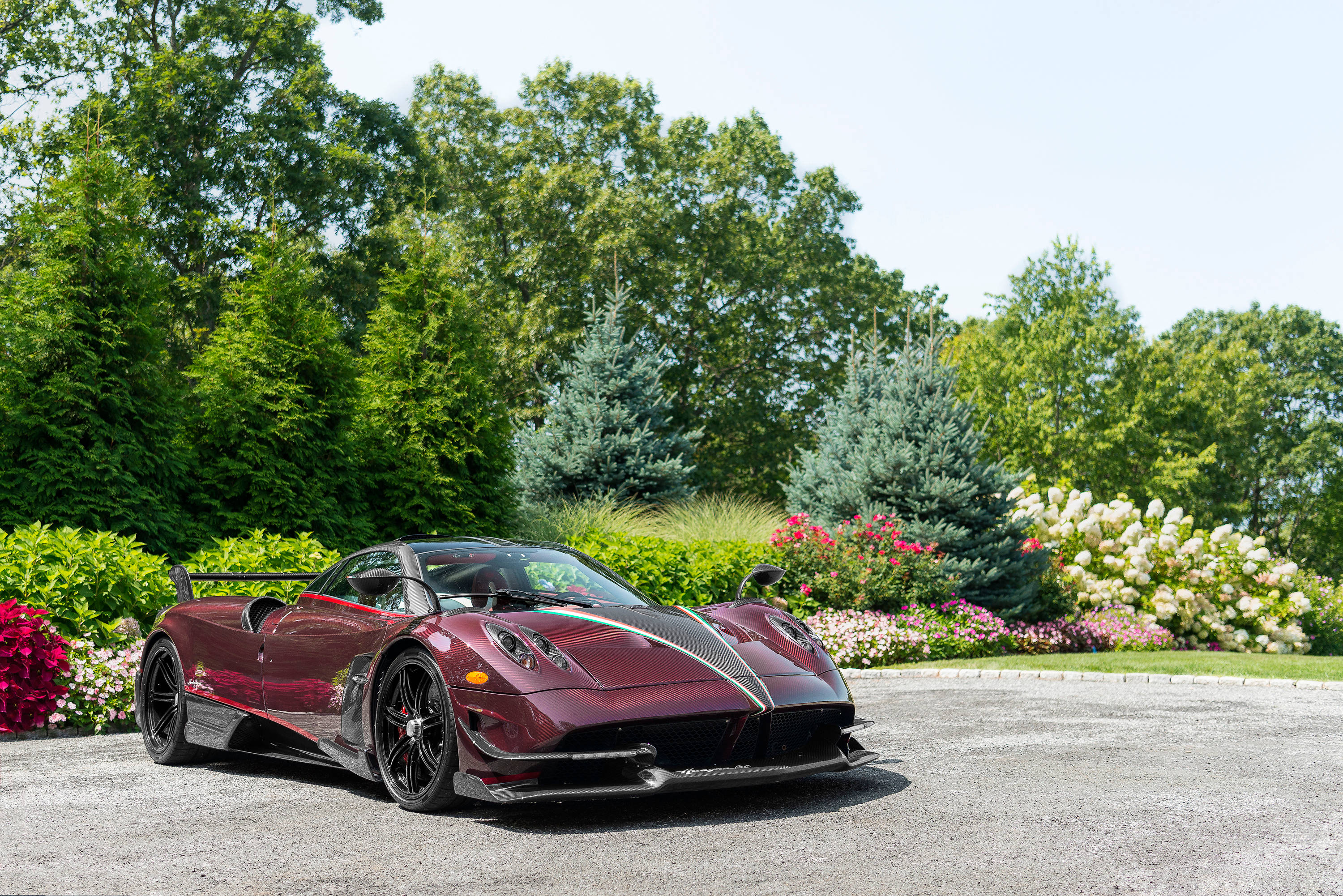 Pagani Huayra BC 紫紅色款 壁紙 — 一輛時尚的栗色帕加尼Huayra跑車在明亮的陽光下閃閃發光，停在碎石車道上，周圍環繞著茂密的綠樹和色彩繽紛的花壇。