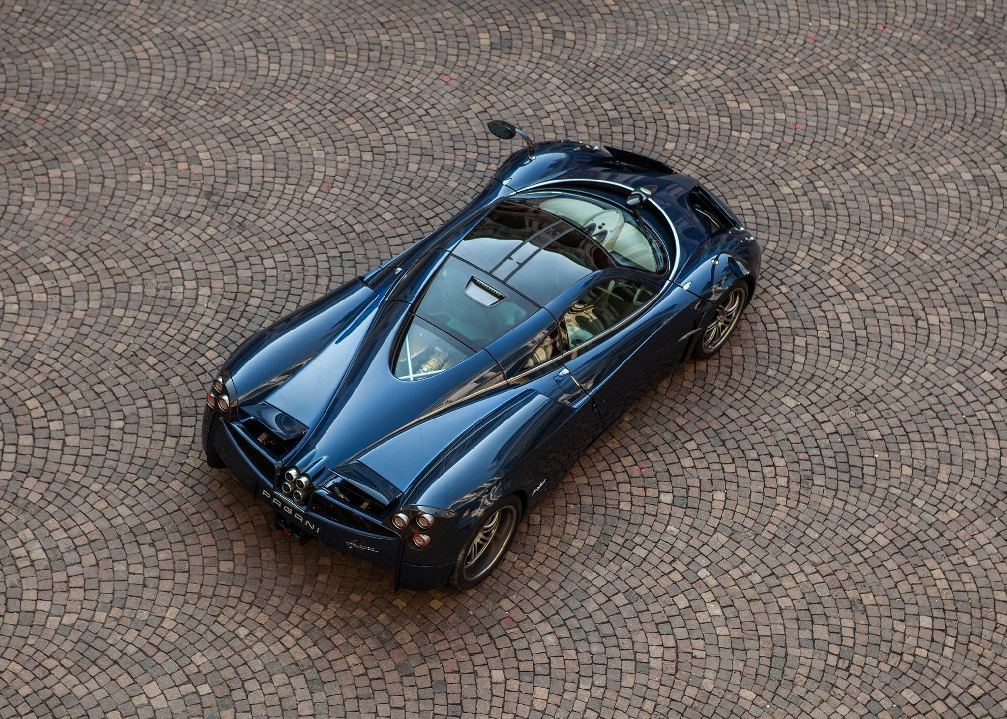Pagani Huayra 深藍色空中俯瞰 壁紙 — 一架流線型藍色Pagani跑車在明亮日光下停泊在彎曲的鵝卵石路面上。