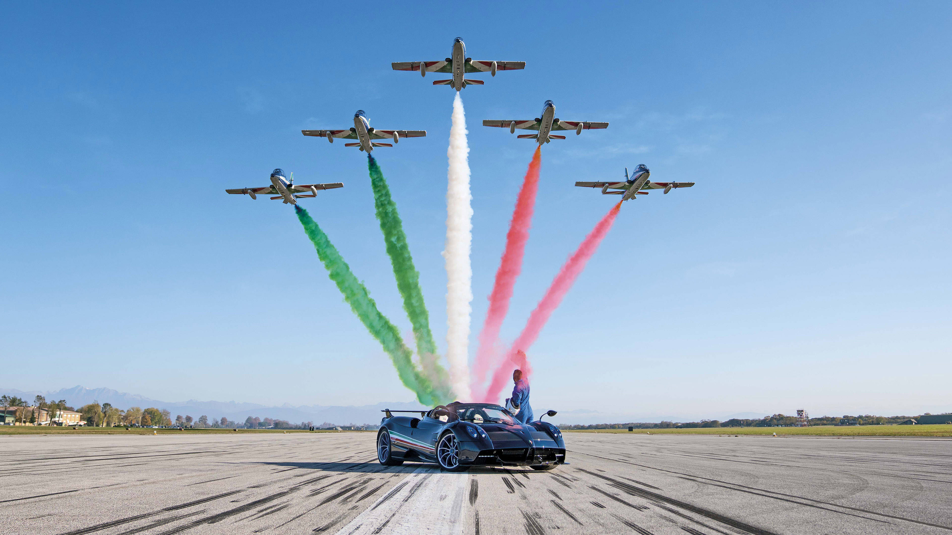 Pagani Huayra 空中表演 壁紙 — 一架時尚的藍色跑車在跑道上，四架噴射機飛過頭頂，留下綠色、白色和紅色的煙霧，形成意大利國旗。