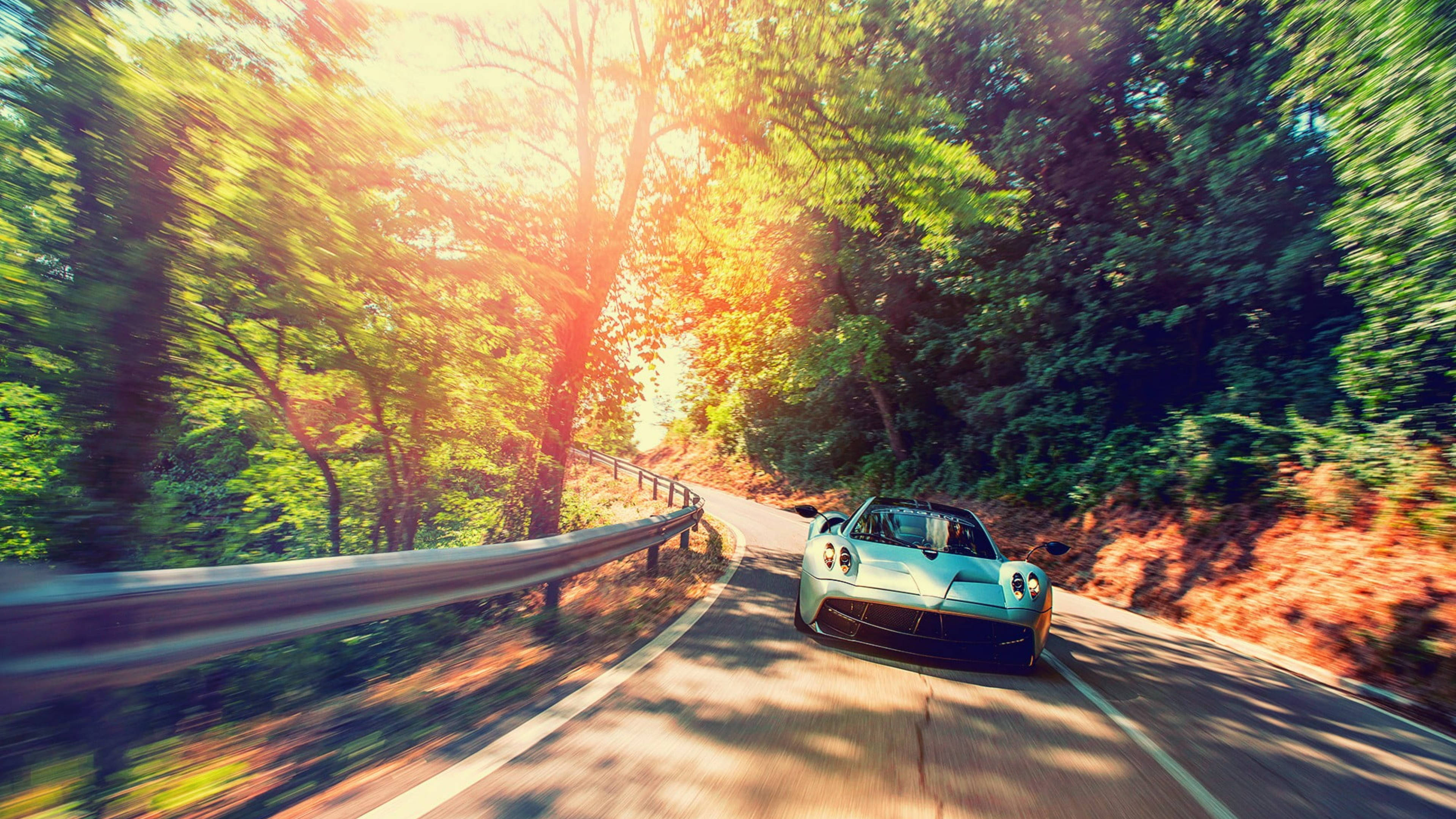 Pagani Huayra 自然動態模糊 壁紙 — 一輛時尚的淺藍色跑車沿著蜿蜒的森林道路飛馳，道路沐浴在透過茂密綠樹灑落的金色陽光中。