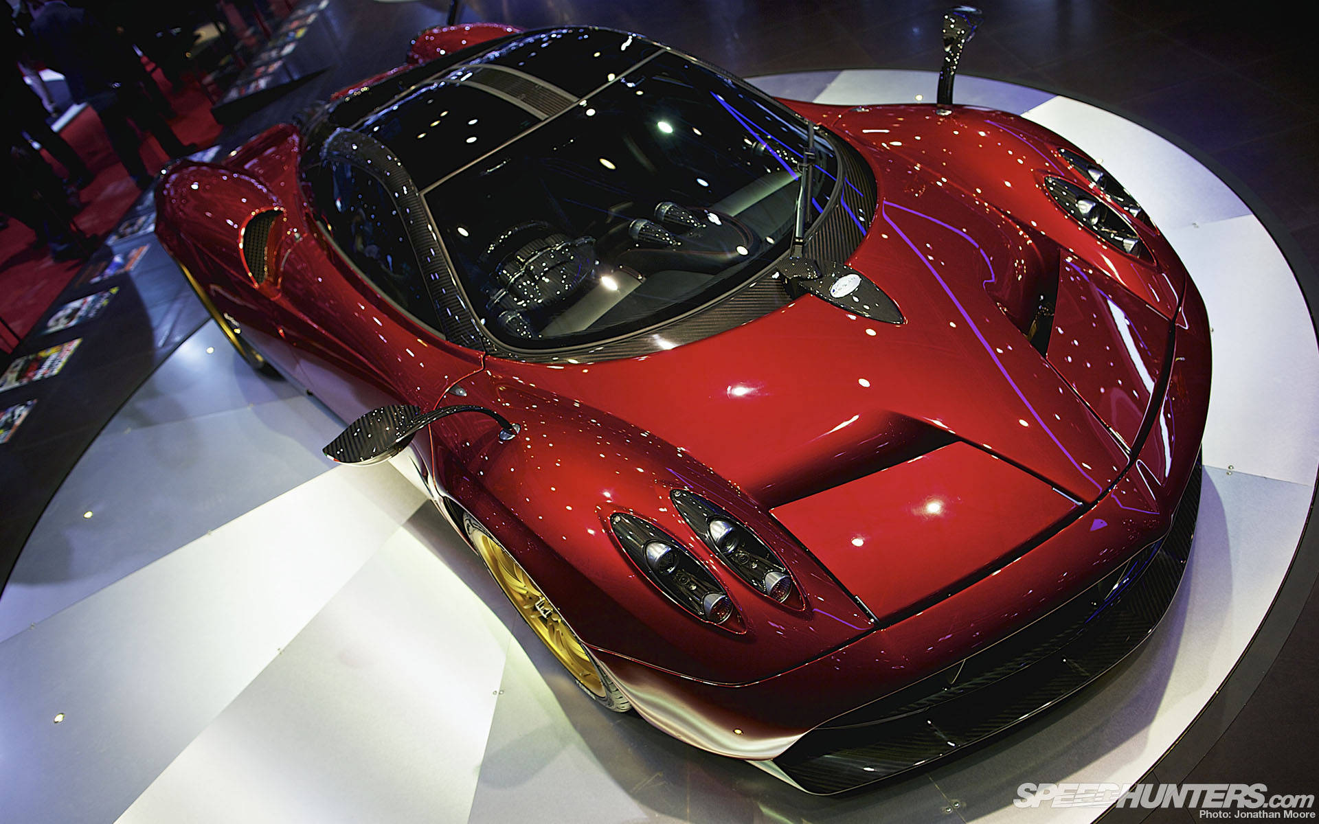 Pagani Huayra 高光紅色超跑 壁紙 — 一輛光澤的紅色帕加尼超級跑車在車展的白色圓形平台上，在明亮的燈光下閃閃發光。