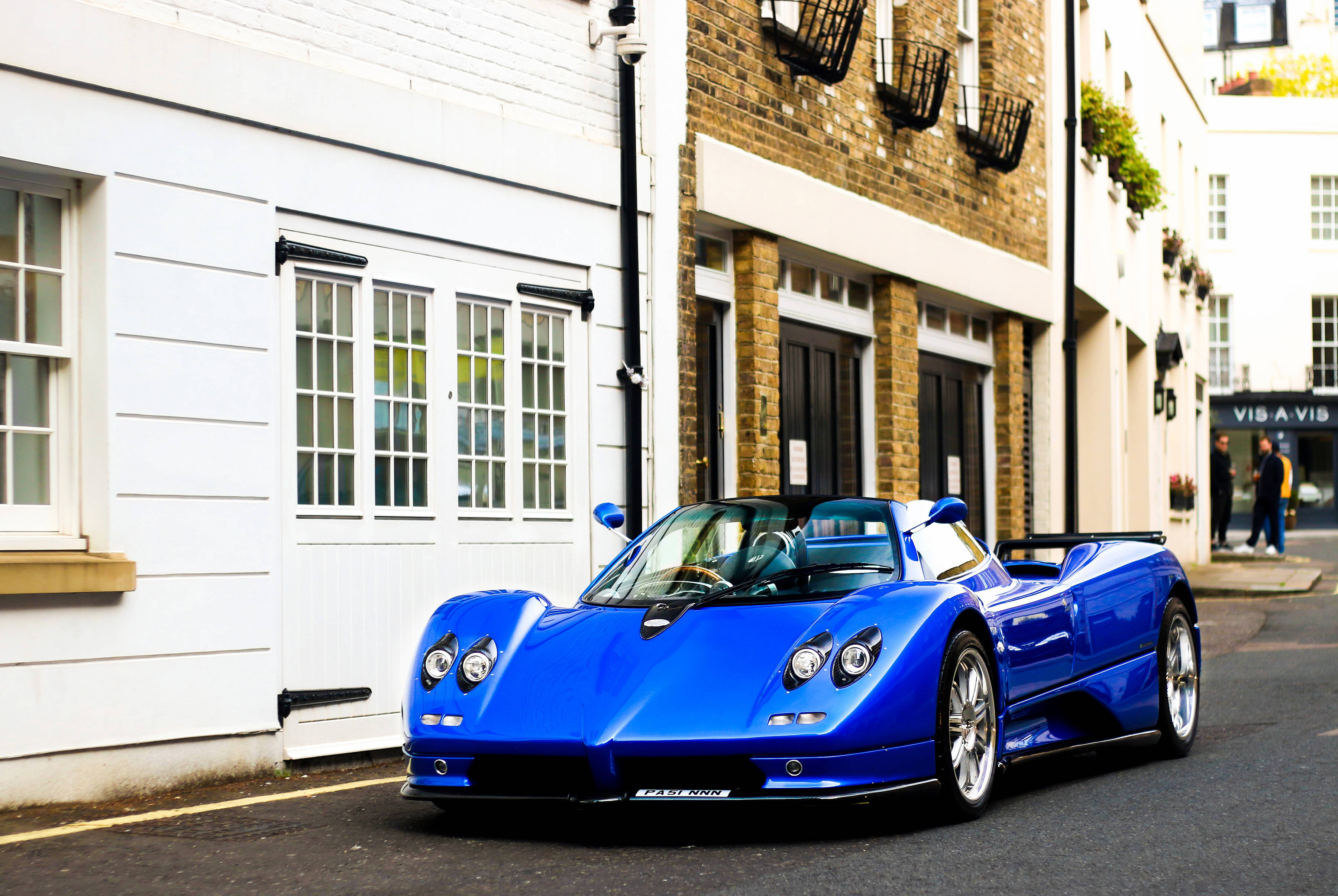皇家藍 Pagani Zonda 壁紙 — 一輛充滿活力的藍色Pagani Zonda跑車停泊在一條狹窄的城市街道上，兩旁是白色和磚砌的建築物。