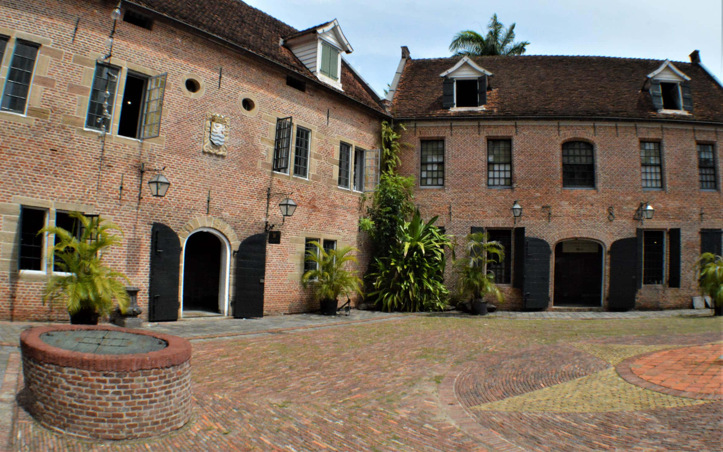 Paramaribo 的蘇里南博物館 壁紙 — 一個歷史悠久的磚砌庭院，設有拱形門口、棕櫚樹和中央噴泉，天空明亮。