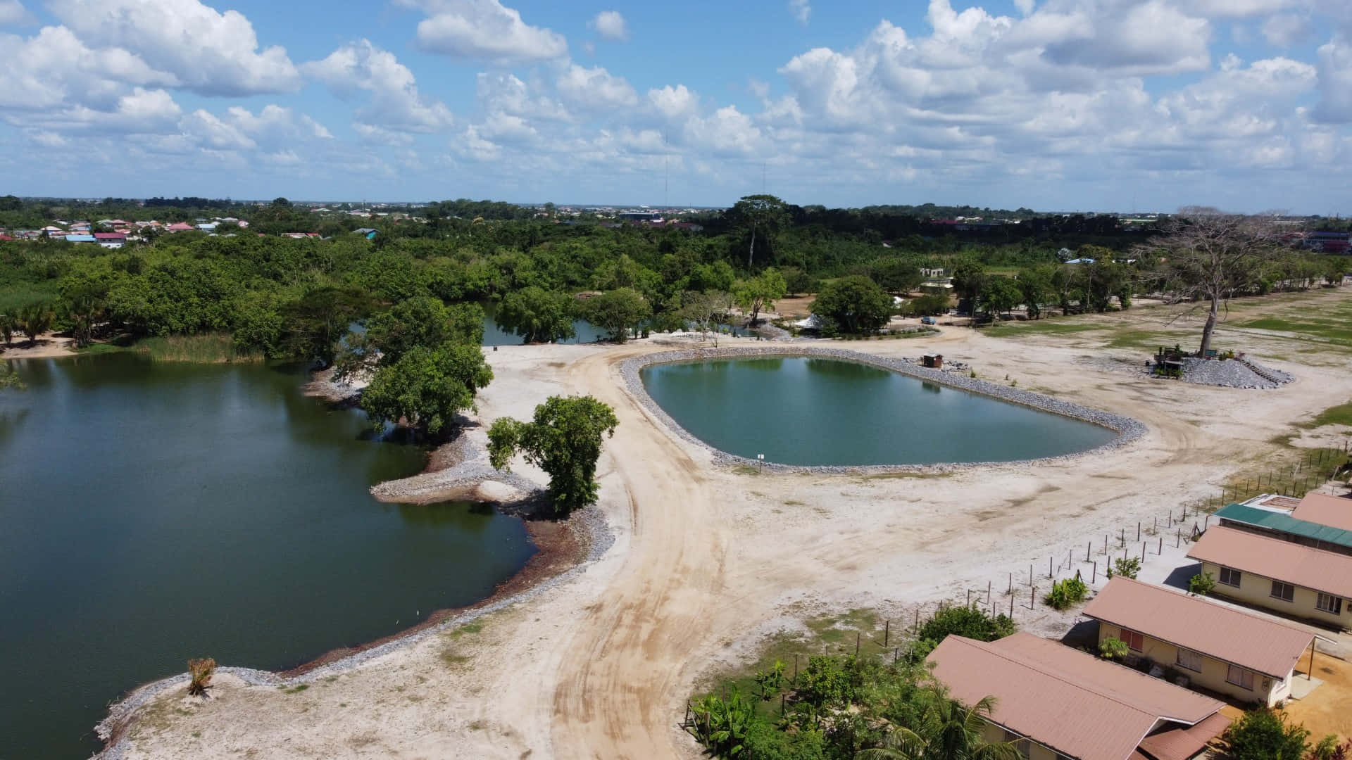 Paramaribo Ring Harbour 項目日落美景 壁紙 — 喺晴朗藍天同 fluffy 雲朵下，一個大型人工池塘嘅航拍圖，周圍係茂密嘅綠樹同泥路。