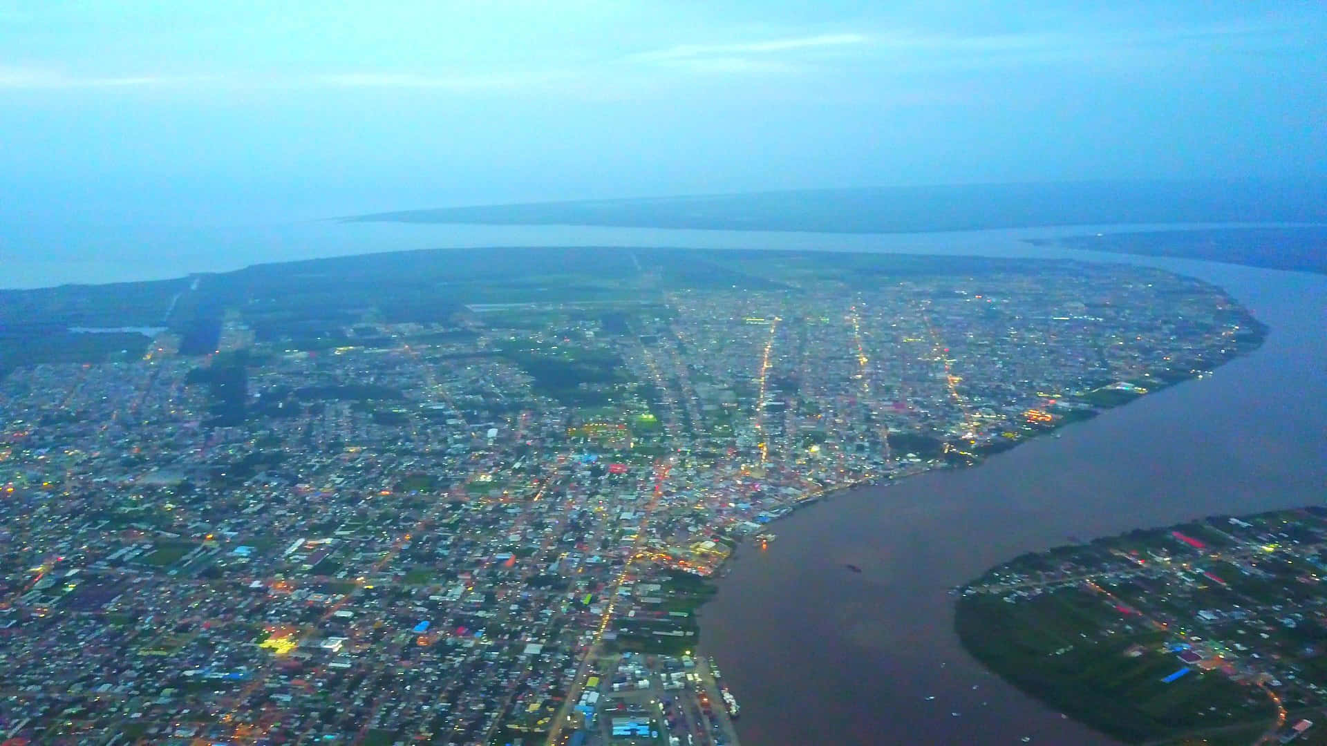 Paramaribo 上空鳥瞰 壁紙 — 俯瞰一座沿著寬闊河流而建的廣闊城市，在暮色中閃爍著街燈和建築物的燈光。