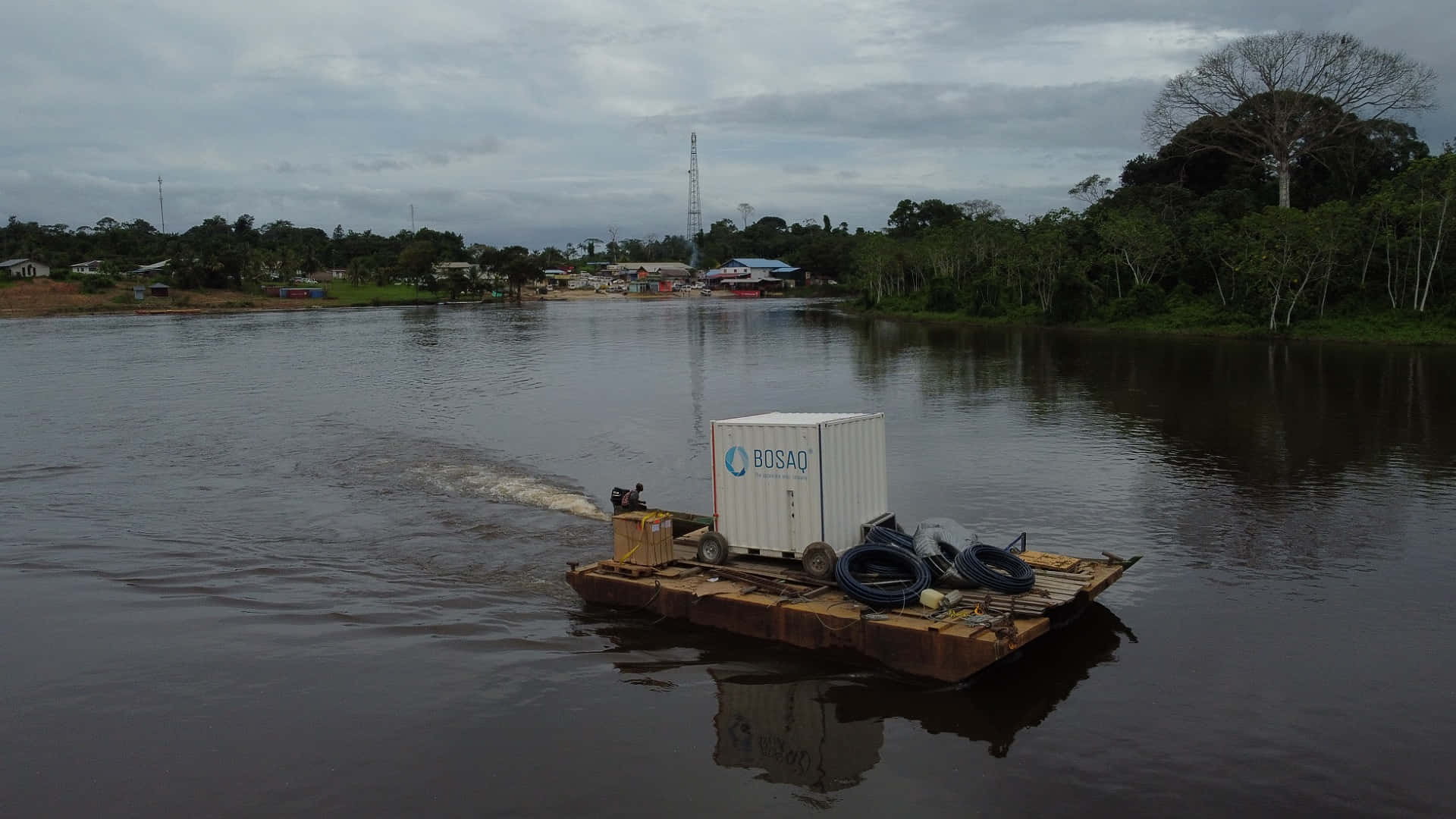 Paramaribo 能源電池於蘇里南河 壁紙 — 一艘破舊的駁船載著一個白色貨運集裝箱和盤繞的軟管，在陰天之下，駛過一條寬闊的河流，遠處有樹木，岸邊有一個小定居點。