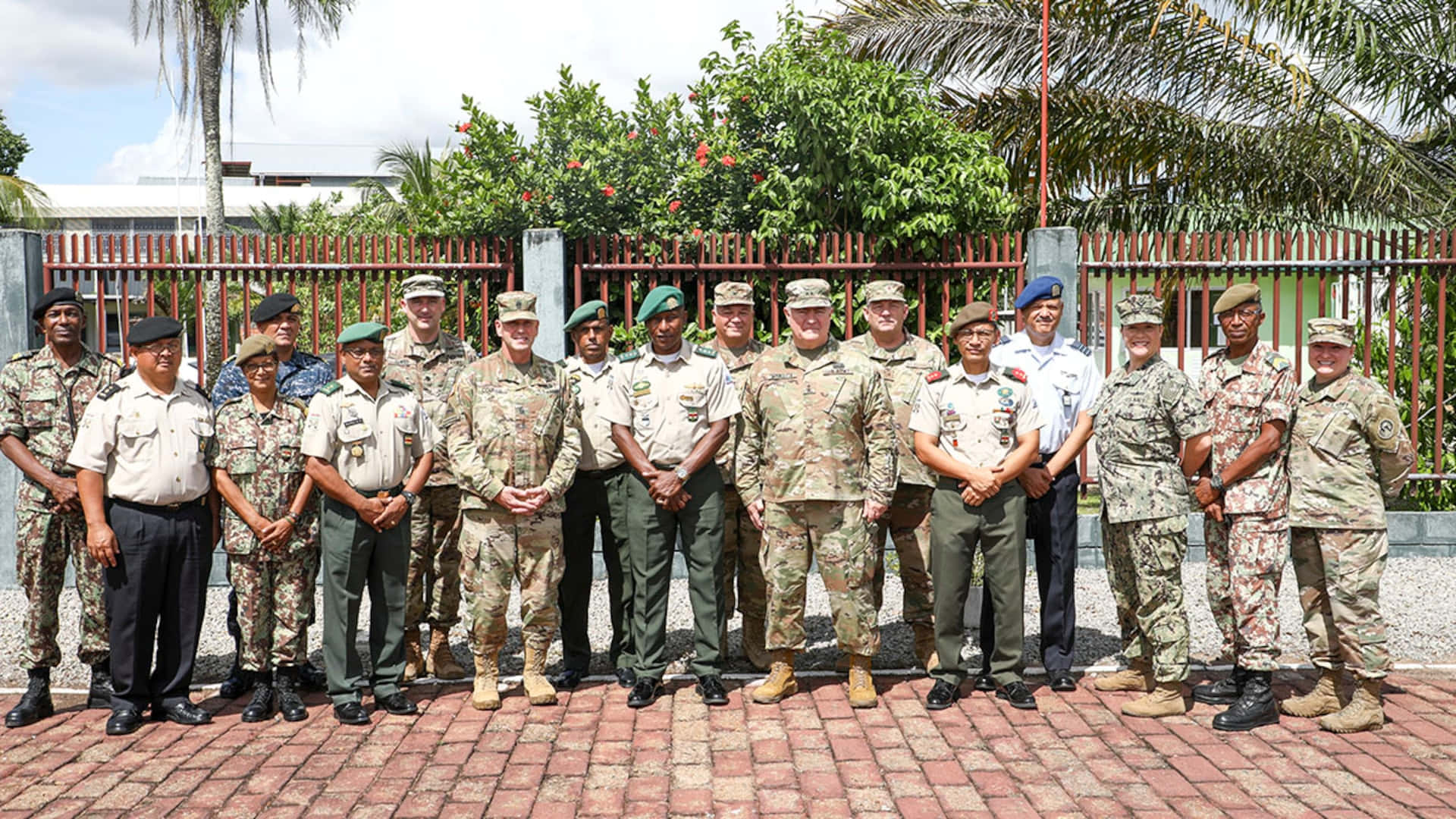 Paramaribo 軍事官員群像 壁紙 — 一群穿著不同制服的多樣化軍官在磚砌庭院中，周圍環繞著熱帶植物和紅色圍欄，為正式照片擺姿勢。