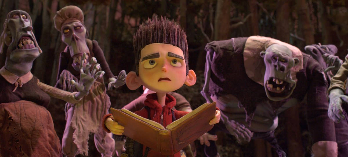 ParaNorman 中 Norman 入迷閱讀魔法書 壁紙 — 一個穿著紅色連帽衫的小男孩在黑暗的森林中，被一群兇猛的殭屍般生物包圍，緊張地讀著一本書。