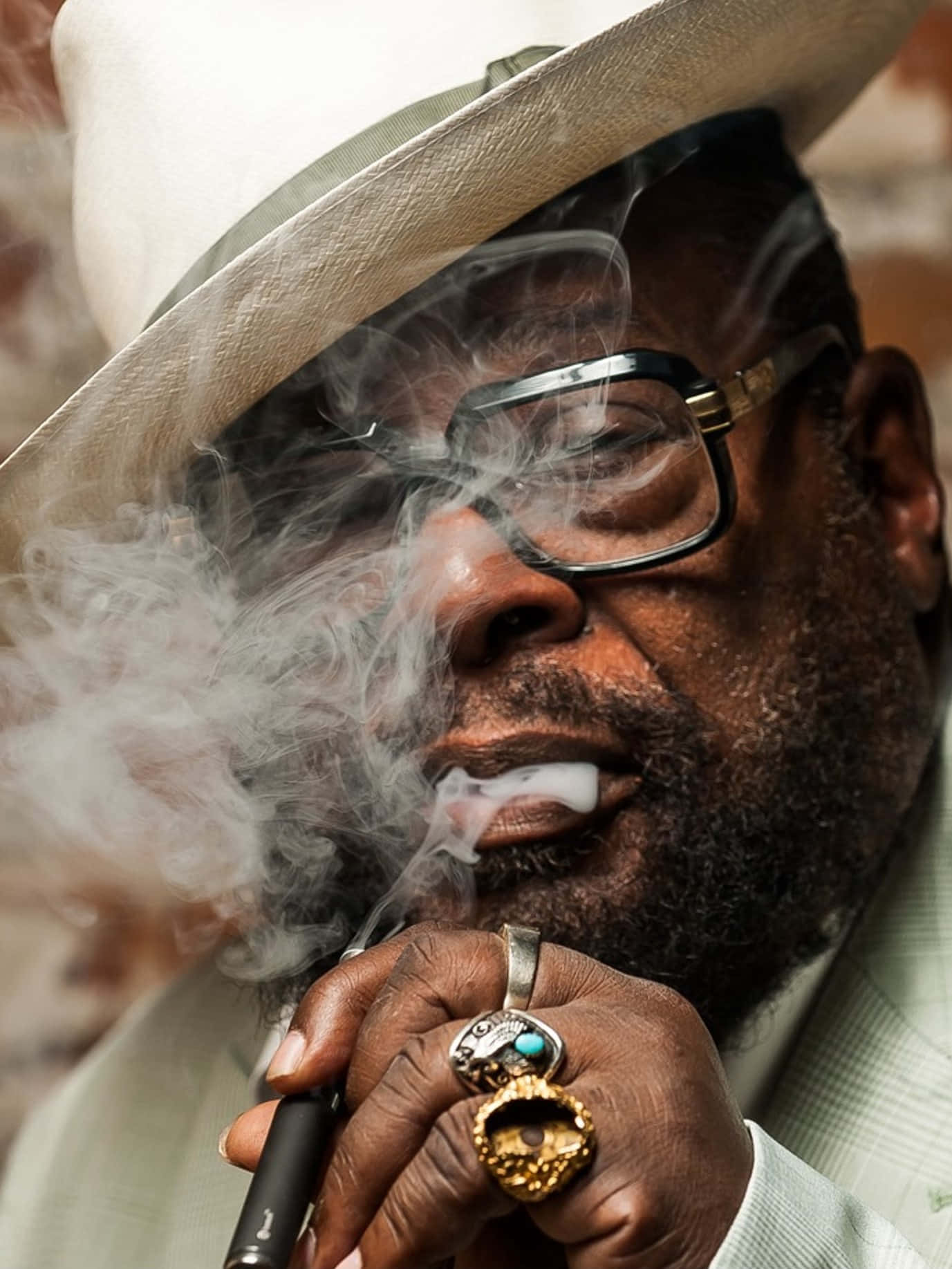 Parliament Funkadelic 乐队的 George Clinton 壁紙 — 一位時尚男士戴著白色帽子和眼鏡，手持電子煙筆呼出煙霧的特寫肖像。