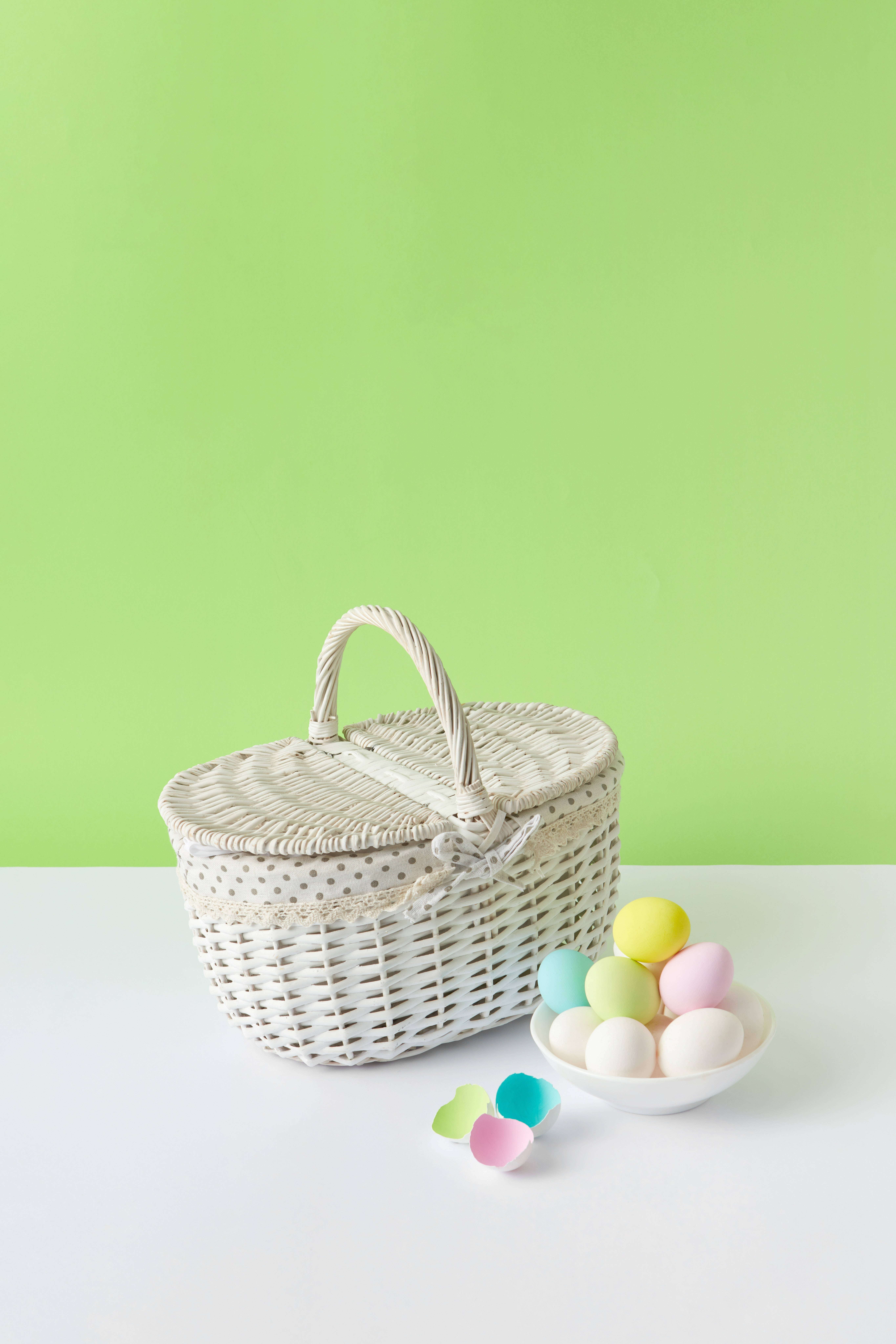 籃子與彩蛋於 Pastel 風綠色背景 壁紙 — 一個白色柳條復活節籃子，帶有圓點飾邊，旁邊放著一碗粉彩雞蛋和散落的蛋殼半邊，背景是明亮的綠色牆壁。