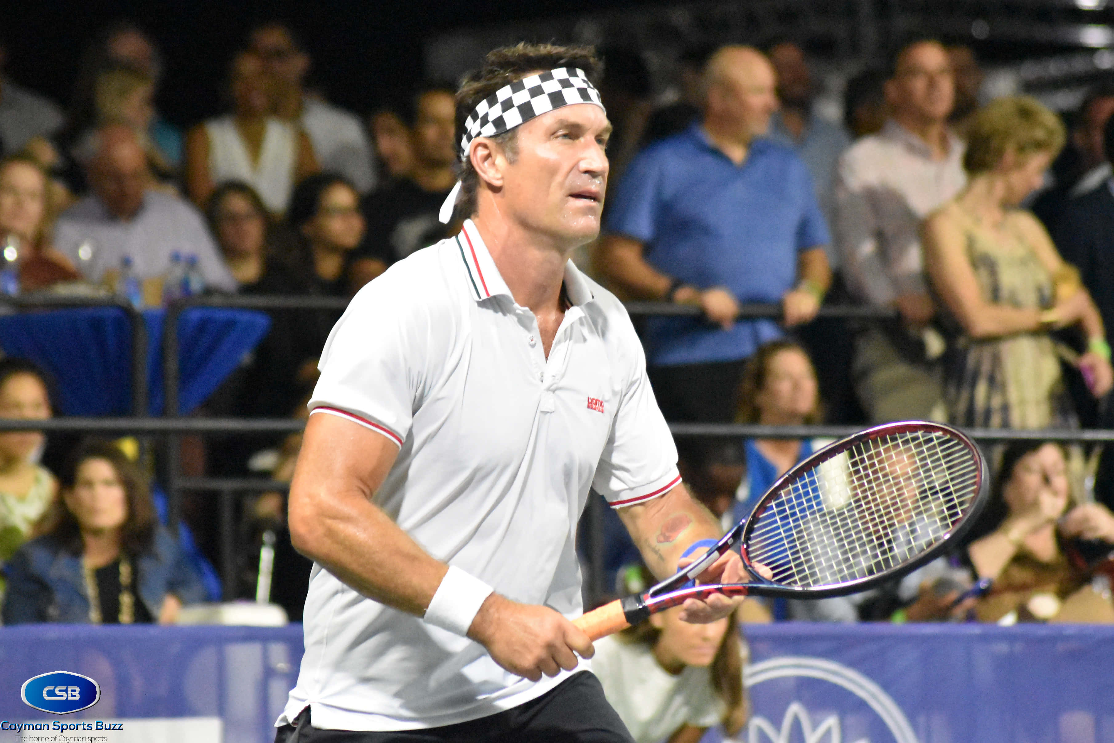 Pat Cash 頭戴髮帶賽場專注照 壁紙 — 一位專注的男性網球運動員，身穿白色襯衫和格子頭帶，在明亮的體育場燈光下於比賽中握緊球拍。