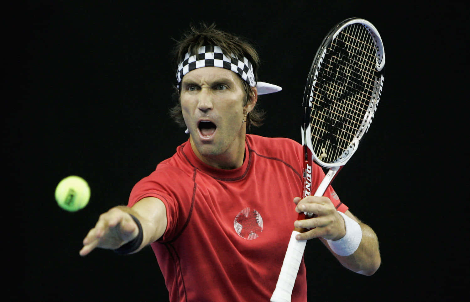 Pat Cash 的攝影焦點 壁紙 — 一名身穿紅色襯衫和格子頭帶的男性網球運動員，在黑色背景下以強烈的專注力向前猛衝，擊打一個亮黃色的網球。