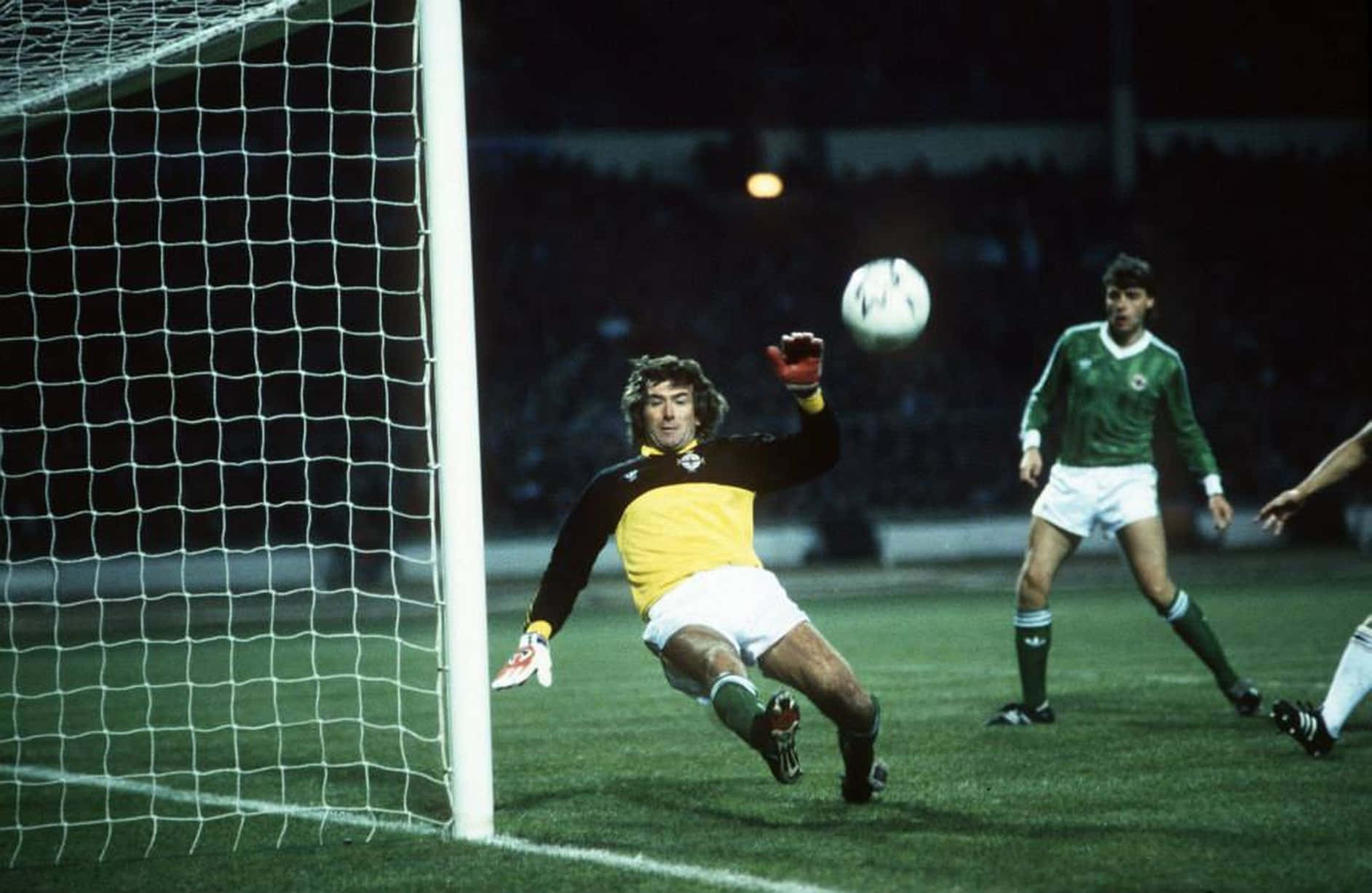 Pat Jennings 參與 FIFA 世界盃小組賽第三組資格賽 壁紙 — 一位身穿黃黑復古足球守門員在球場燈光下飛身撲向球門網附近的球。
