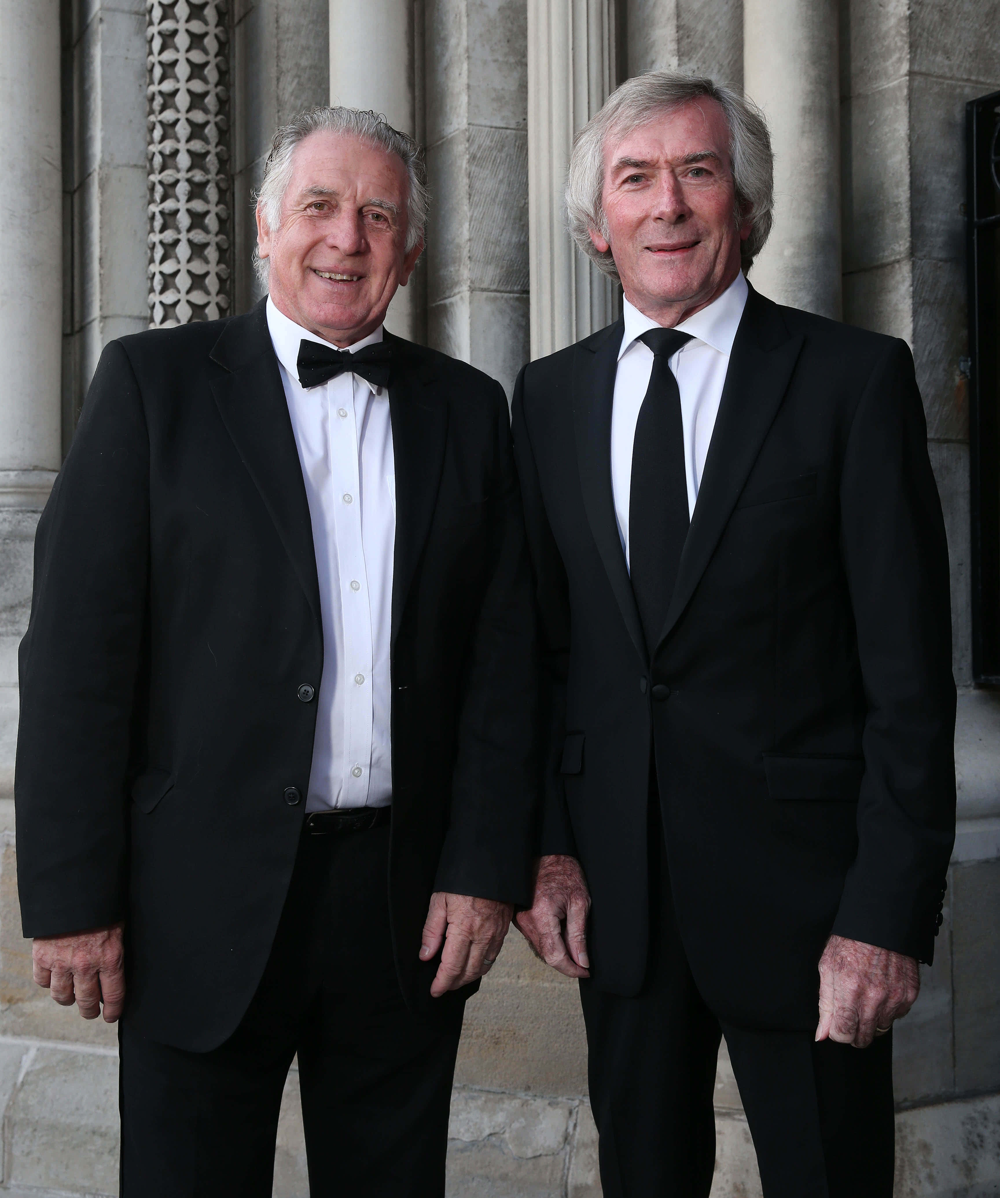 Pat Jennings 與 Gerry Armstrong 於 2019 年 Ulster Tatler 獎典禮 壁紙 — 兩位微笑的老年男士身穿正式黑色西裝，並排站在帶有華麗柱子的石建築外牆前。