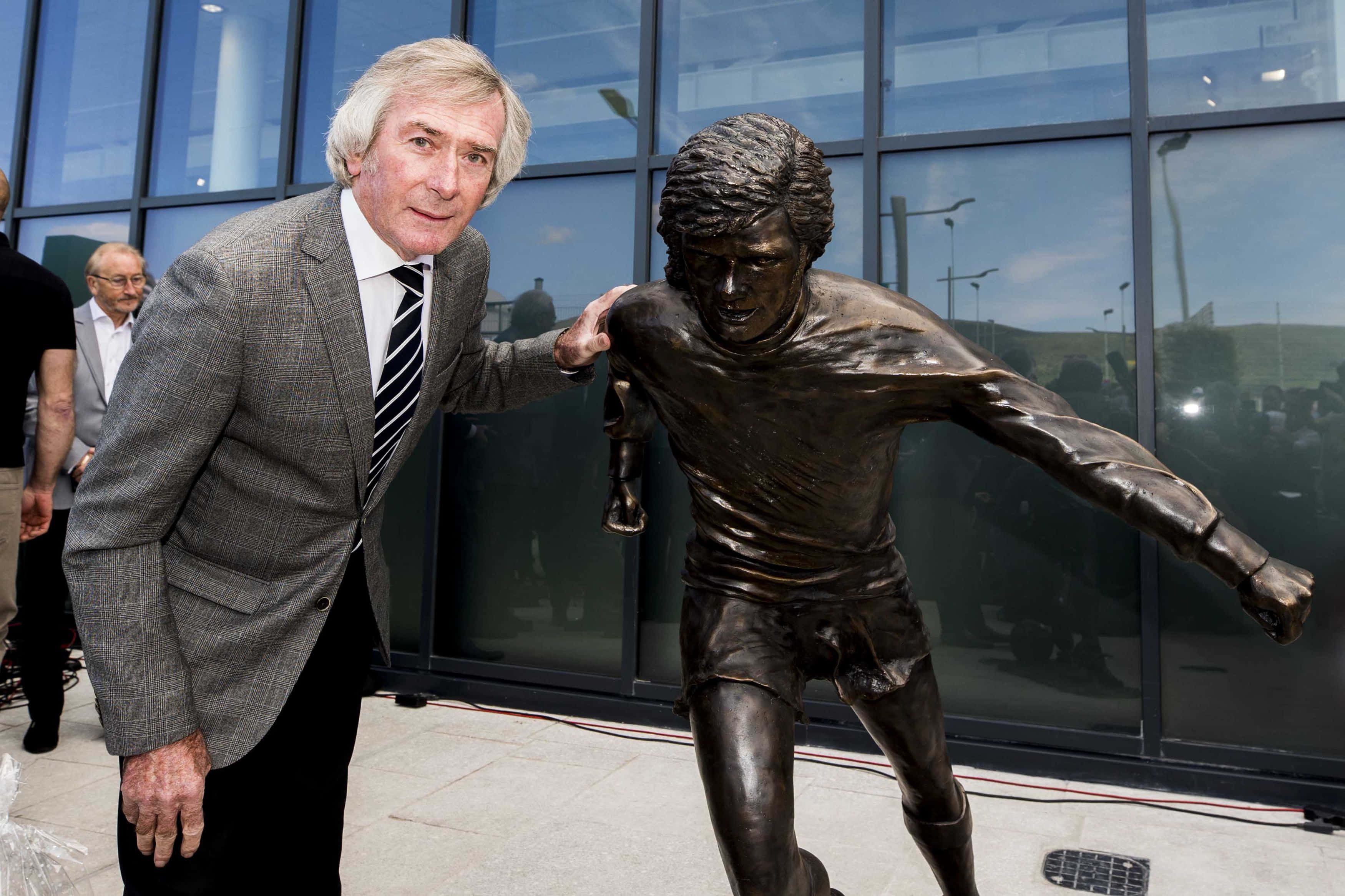 Pat Jennings 與曼聯傳奇 George Best 的雕像於貝爾法斯特 壁紙 — 一位穿著西裝的年長男士輕輕將手放在一個動感十足的足球員青銅雕像上，雕像呈現大步奔跑的姿態。