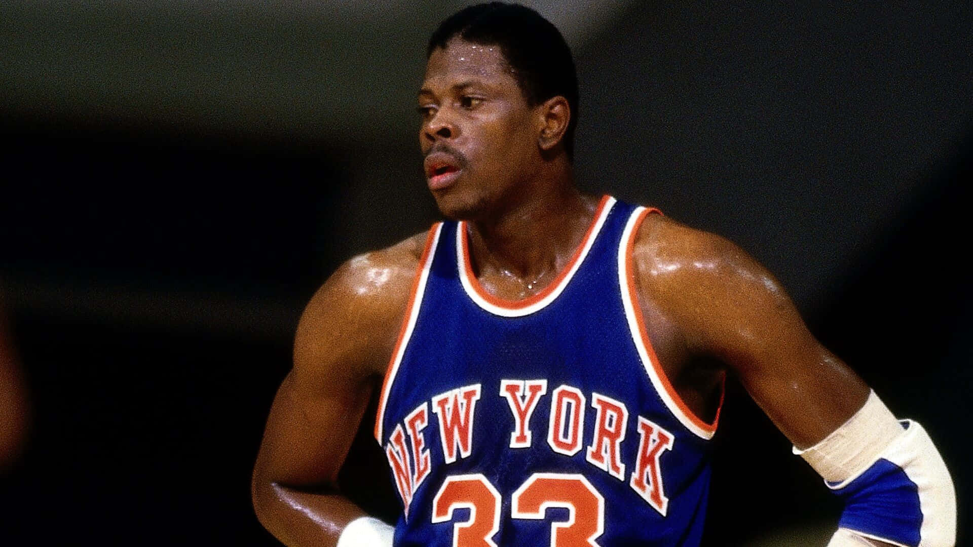 帕特里克·尤因籃球中鋒NBA球員 壁紙 — 一名汗流浹背的籃球運動員身穿紐約人球衣，在明亮的場館燈光下比賽中凝視著。