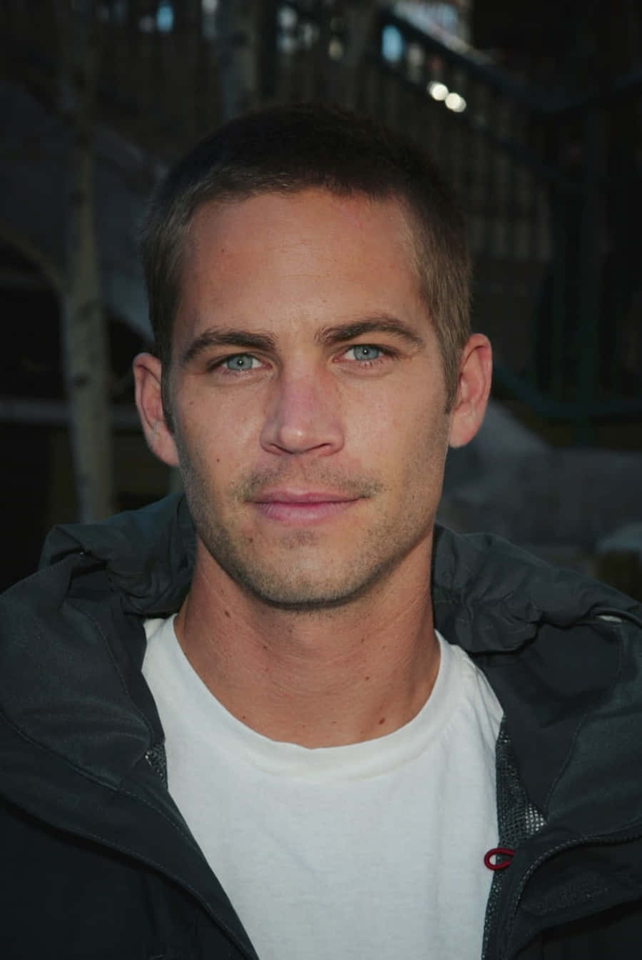 保羅·獲加 桌布 壁紙 — Paul Walker 嘅特寫肖像，佢有對強烈嘅藍眼睛，著住一件深色外套喺白色T恤外面，背景係模糊嘅城市景觀。
