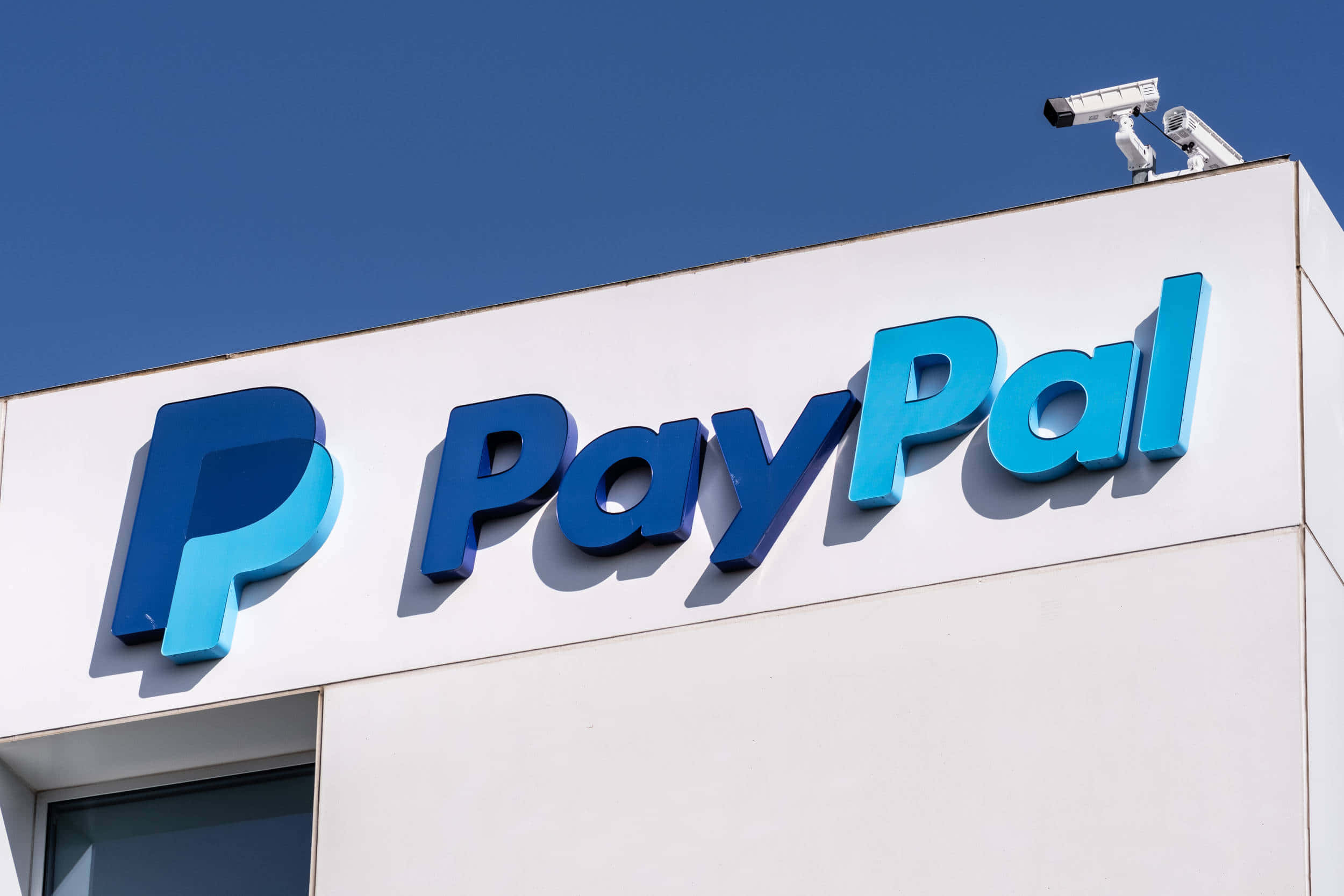 透過 PayPal 輕鬆發送與接收資金 壁紙 — 鮮明的藍色PayPal標誌安裝在白色建築物上，上方有藍天和保安攝像頭。