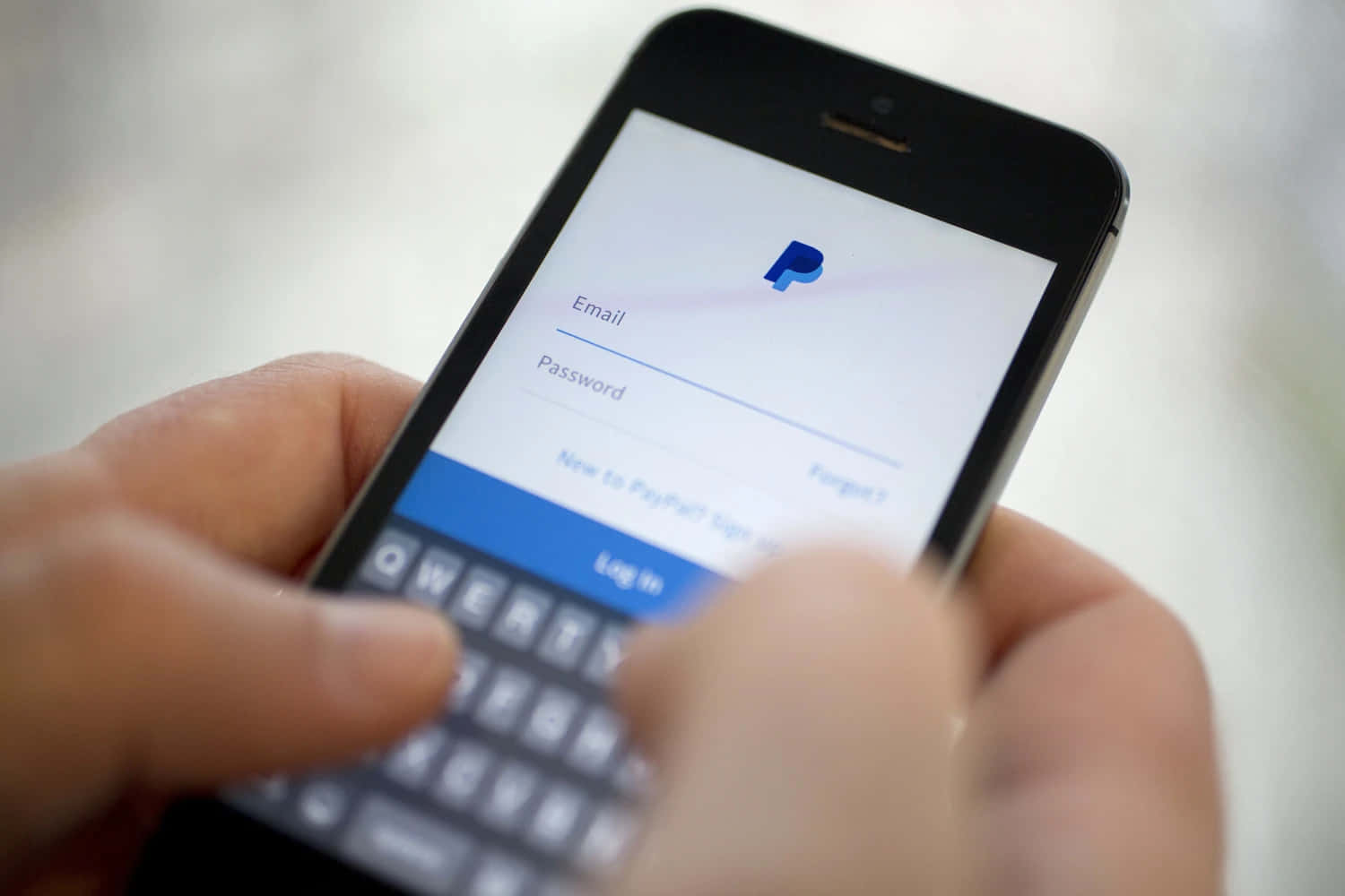 透過 PayPal 輕鬆完成付款與收款 壁紙 — 特寫鏡頭：雙手在模糊背景的智能手機上輸入PayPal應用程式的登入憑證。