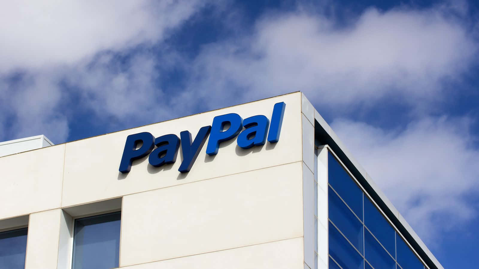 透過 PayPal 安全轉移資金 壁紙 — 一個低角度拍攝的現代建築，帶有大型藍色PayPal標誌，背景是明亮的藍天和蓬鬆的白雲。